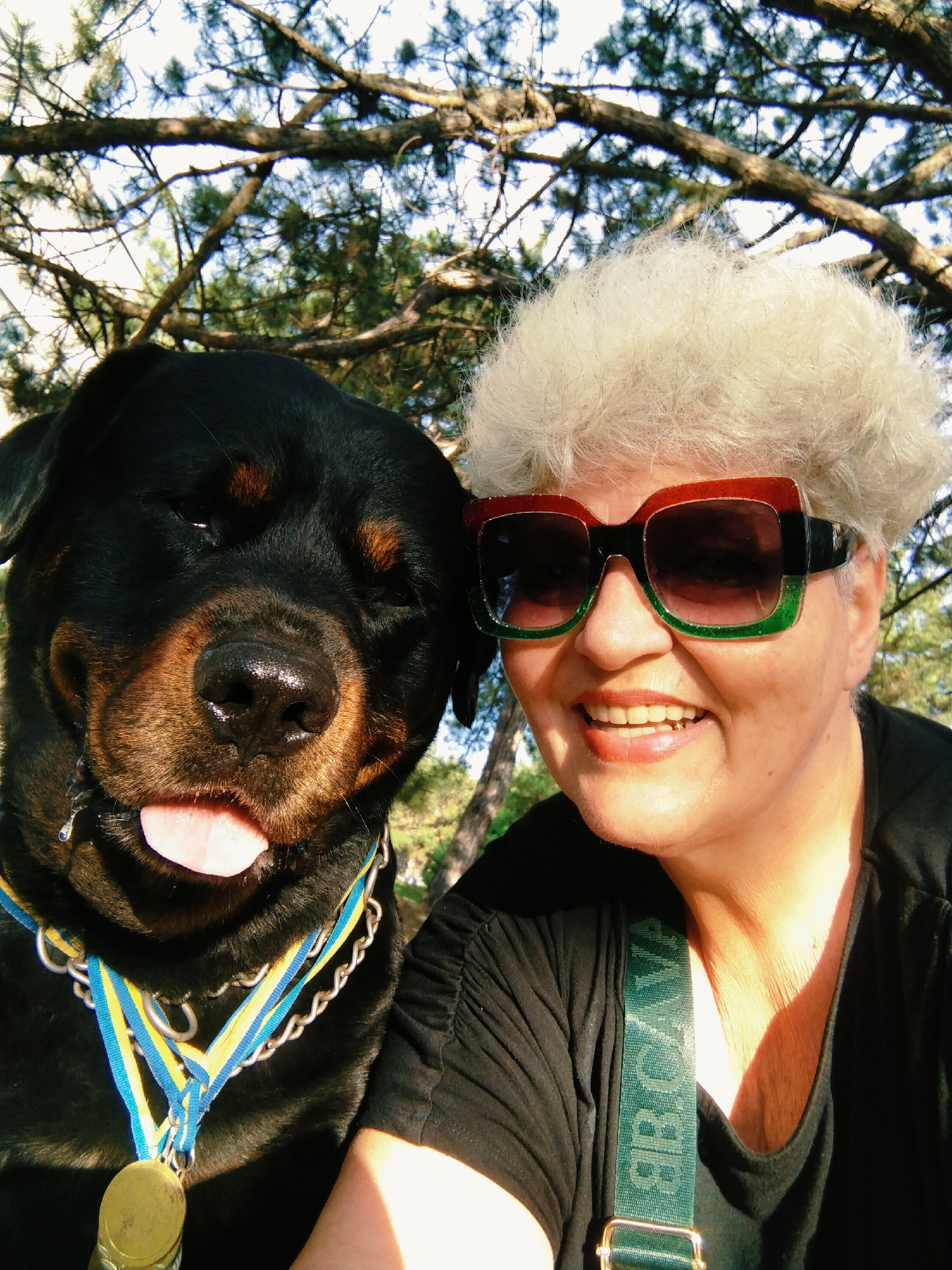 A selfie is taken outdoors, with a Rottweiler and a woman in the foreground. The dog, on the left, is a large black dog with brown markings around its eyes and muzzle. It is wearing a blue and yellow medal with a chain collar. The dog's tongue is hanging out, and it has a droopy expression. The woman, on the right, has fair skin and short, fluffy white hair. She is wearing large, colorful sunglasses with red and green rims and a black top. A green strap with white lettering is visible across her shoulder. Both the dog and the woman appear to be looking at the camera. The background consists of sun-dappled tree branches and foliage, suggesting an outdoor setting in a park or wooded area within the city of Horishni Plavni, Ukraine. The lighting indicates it is daytime, likely sunny.