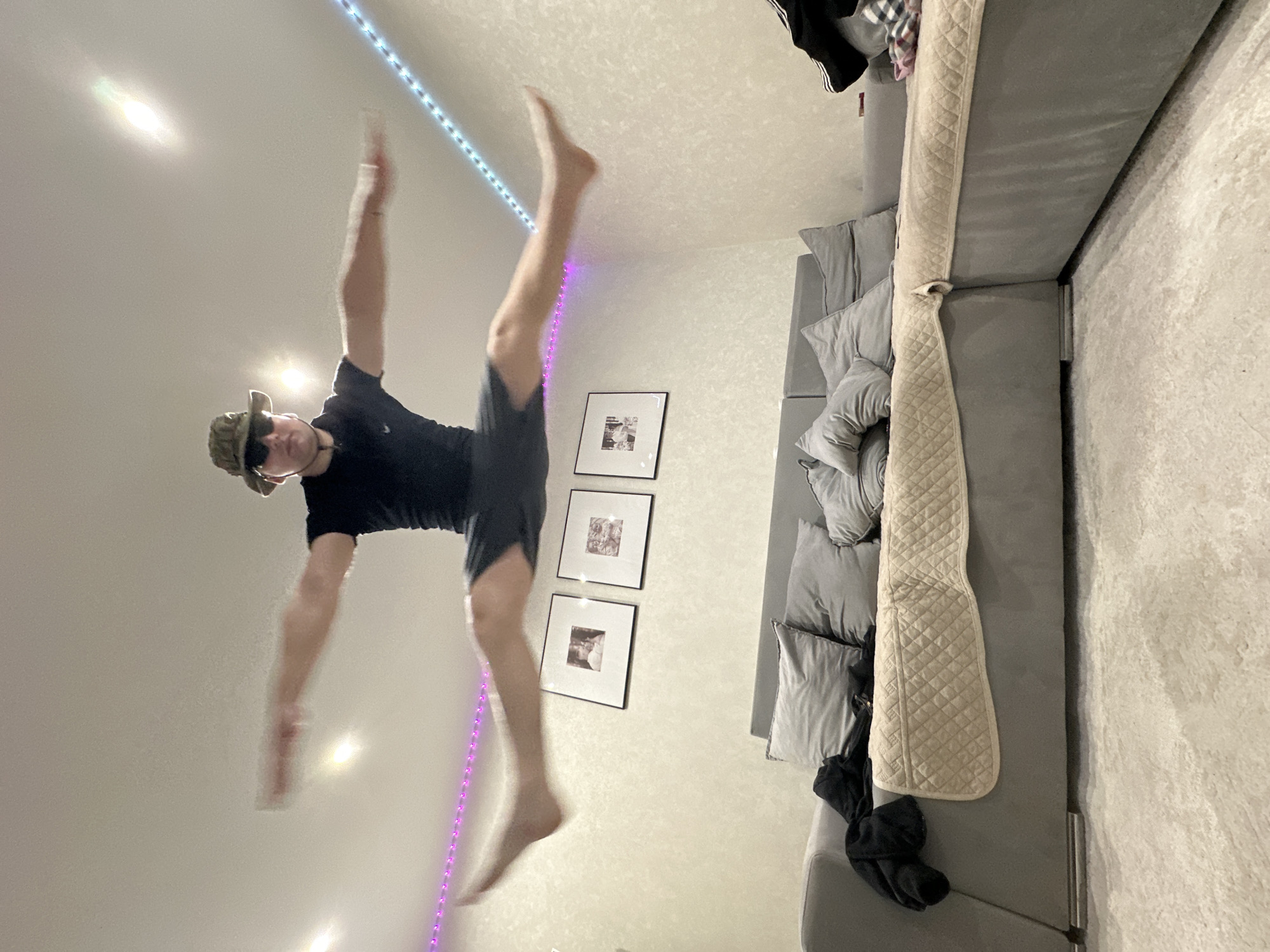 A person is in mid-air in what appears to be a living room. The person is wearing a black shirt, dark shorts, a hat, and sunglasses. Their arms and legs are extended. There is a couch with pillows along the right side of the image. On the wall, there are three framed pictures and an LED light strip. The exact location in Chaikovsky, Russia, cannot be determined from the image.