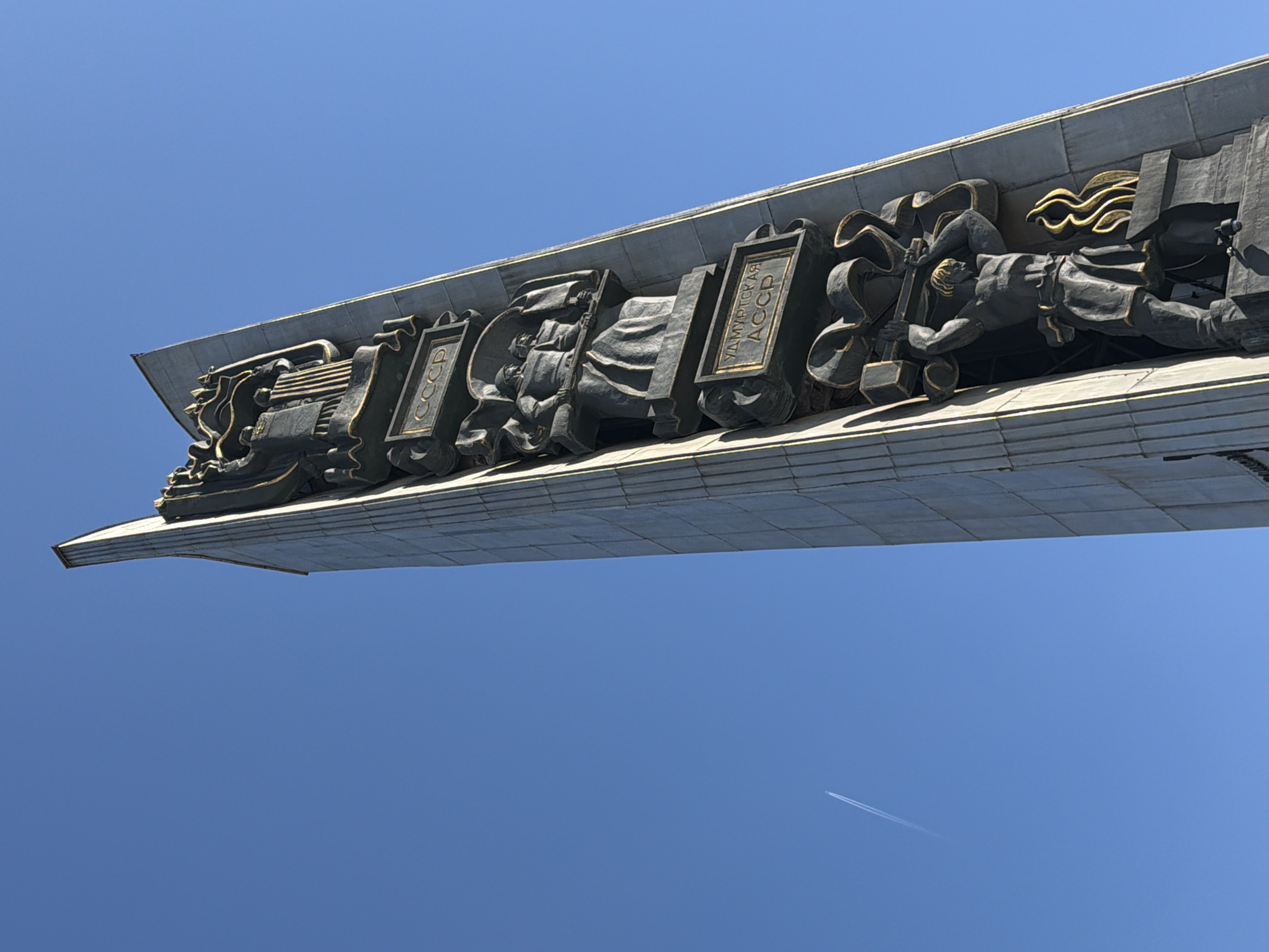 A tall, tapering concrete stele, identified as the Monument to the Friendship of Peoples, is depicted in Izhevsk, Udmurt Republic, Russia, located on Central Square. The monument features multiple deeply recessed bronze bas-reliefs with gold-painted accents. Visible elements, from top to bottom, include: an upper panel depicting figures with banners above a rectangular plaque inscribed with "CCCP" (USSR); a central panel showing two figures, one appearing as a soldier with a rifle and the other a worker, positioned above a plaque bearing the inscription "УДМУРТСКАЯ АССР" (Udmurt Autonomous Soviet Socialist Republic); and a lower panel featuring a muscular male figure, likely a blacksmith, wielding a large hammer over an anvil, with stylized gold flames to his right. The stele's angular, sharp-edged apex is visible. The scene is set against a clear, bright blue sky, with the diagonal contrail of a distant aircraft present in the upper-left portion of the frame. Strong sunlight illuminates the monument, creating distinct shadows within the recesses and on its surfaces. No human subjects are visible in the immediate vicinity or at the base of the monument within the frame.
