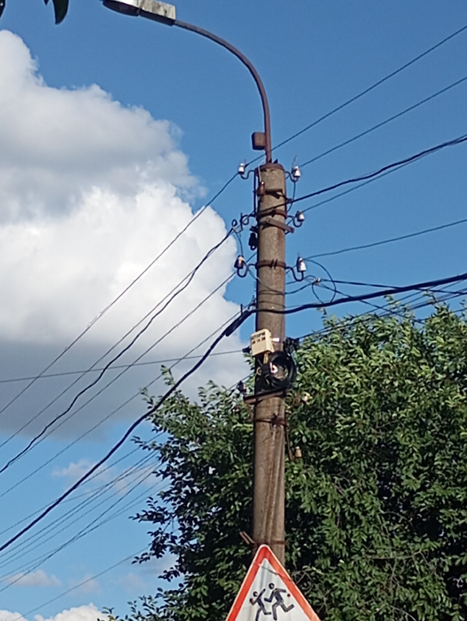 A concrete utility pole stands against a bright blue sky with scattered white clouds and is surrounded by lush green foliage. Multiple dark electrical wires stretch horizontally across the frame, supported by ceramic insulators attached to the pole. A street lamp extends from the top of the pole towards the left. On the pole, a small, light-colored electrical box is mounted, with coiled black cables attached. At the bottom of the image, a triangular red and white sign depicts two black silhouettes of children running, indicating a children crossing or school zone. The overall scene is outdoors, under daylight conditions, and appears to be in an urban or semi-urban environment.