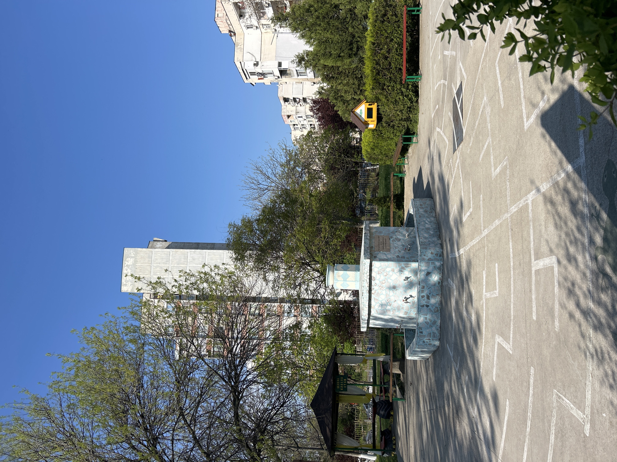 The image shows a playground in Plovdiv, Bulgaria, with painted white lines on a concrete surface forming a maze. The playground features a drinking fountain with a tiled facade, a small gazebo-like structure with a dark roof, and some playground equipment including a small yellow structure. The backdrop includes buildings with white and gray facades, partially obscured by trees. The sky is clear and blue, indicating a sunny day.