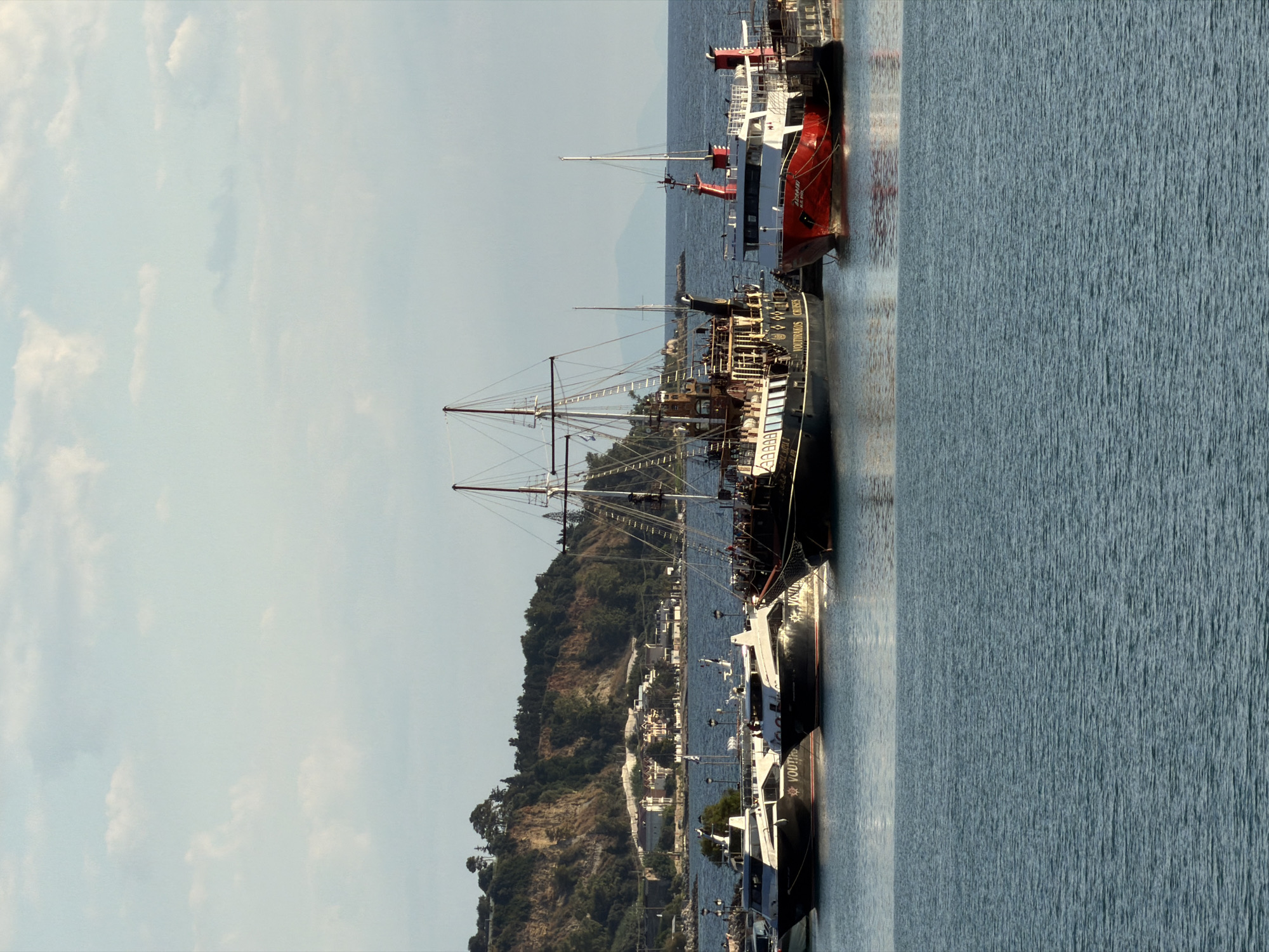 The image shows a harbor in the Zakynthos Municipal Unit, Greece, with several boats docked in the water. In the foreground, the blue water is calm with gentle ripples. In the midground, multiple boats are visible. On the left, two sleek, dark-hulled passenger ferries are docked side-by-side, with "VOUTIRAKOS CRUISES" visible on their sides. To their right, a larger, older-style ship with multiple masts, resembling a pirate ship, is docked. This ship has intricate wooden detailing and is decorated with flags. To the right of the pirate ship, a red and white passenger ferry is docked. In the background, a hilly coastline is covered with trees and dotted with buildings. The sky above is a pale blue with scattered white clouds. The lighting suggests it is daytime, likely late afternoon given the angle of the sunlight.