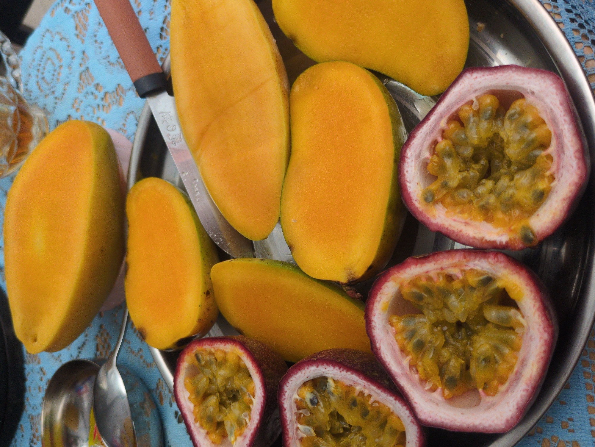 A close-up shows a silver tray filled with cut mangoes and halved passion fruits. A knife rests among the mangoes. A spoon is visible beside the mangoes. The tray sits on a light blue tablecloth with a lace-like pattern. Part of a glass is visible on the left. The background hints at an outdoor setting. Based on the prompt's request, it is inferred the location is the South Quy Nhon Ward, Vietnam.