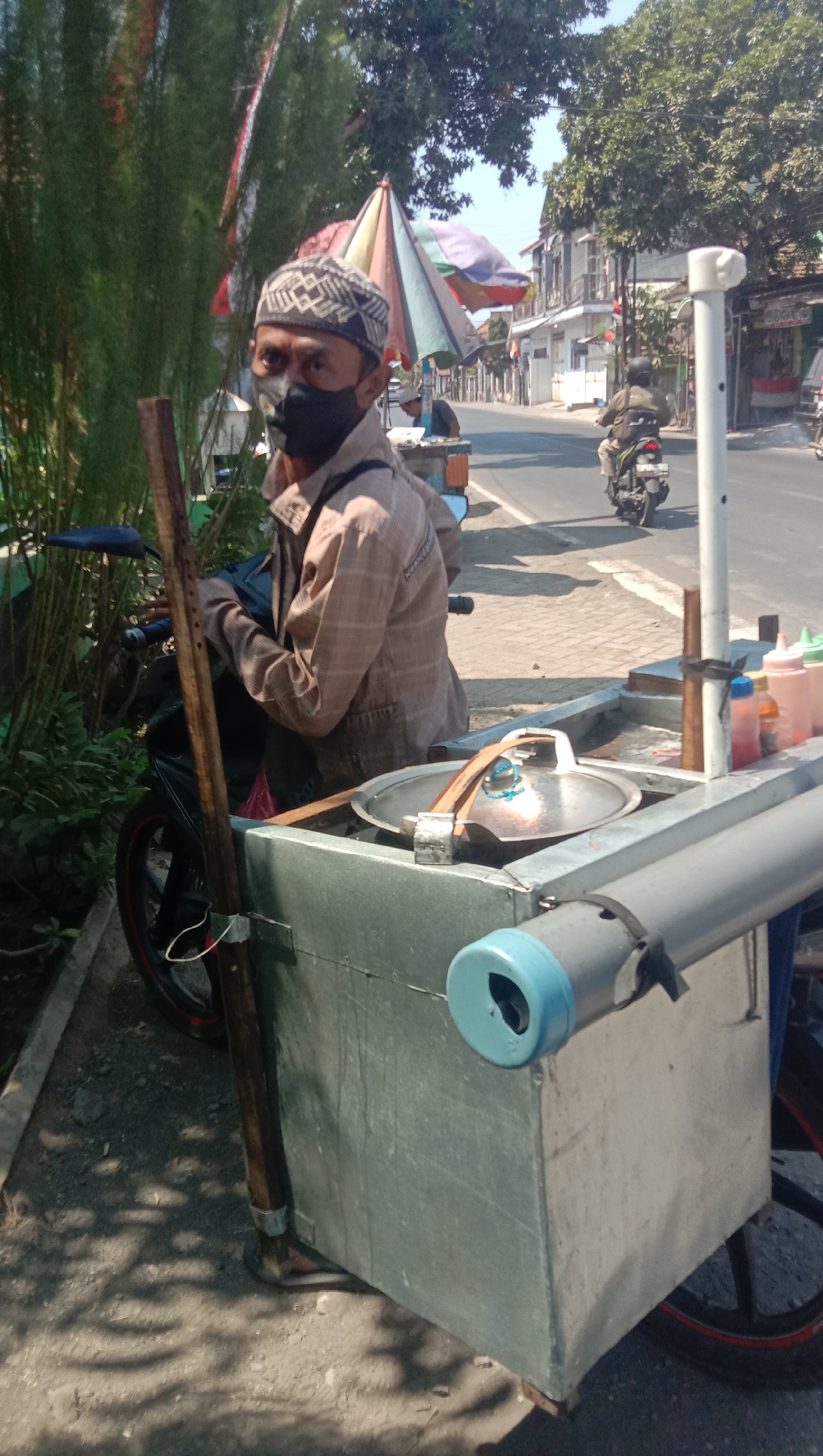A man wearing a patterned skullcap and a face mask is seated on a motorcycle next to a food cart. The cart is made of metal and has wheels. It contains a metal pot with a lid, bottles of condiments, and other items. The man is wearing a light brown plaid shirt. Behind the man, a street is visible with buildings and trees. A motorcycle is passing by on the street. The scene appears to be in an outdoor urban environment.