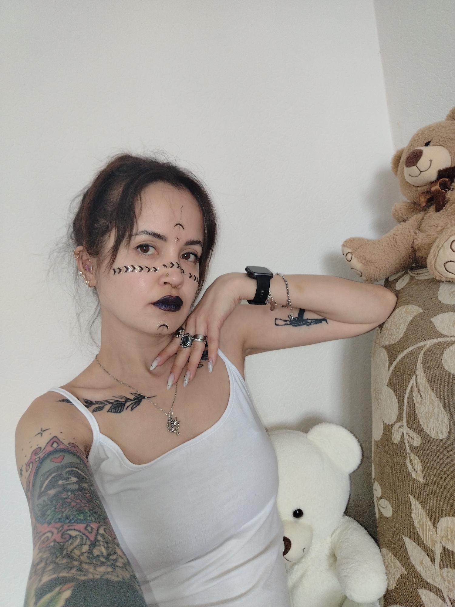 A fair-skinned woman is positioned in front of a white wall and part of a patterned brown and white surface. She is wearing a white tank top and has her hair pulled back. Her left arm, which is heavily tattooed, is in the foreground. Her right arm is bent, resting on the patterned surface, with a smartwatch on her wrist. She has dark makeup on her lips and facial markings consisting of moons and arrows. She is also wearing jewelry, including rings and a necklace.
Two teddy bears are present in the scene. A brown teddy bear is situated on top of the patterned surface, while a white teddy bear is partially visible behind the woman.
There is no information available to determine if the image was taken in Noviy, Russia.
