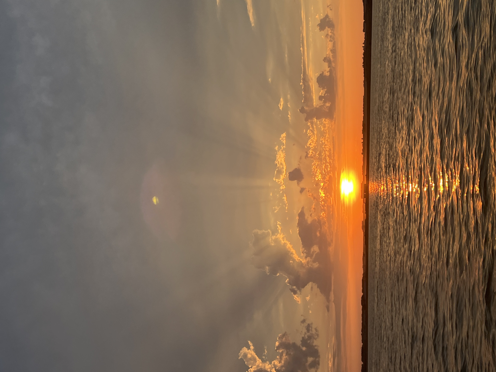 The image depicts a sunset over a body of water, likely a river or lake, with a distant shoreline visible. The sun, a bright orange disc, is low on the horizon, casting a warm glow across the scene. Rays of light emanate from the sun, piercing through scattered clouds that are tinged with orange and yellow hues. The water's surface is rippled, reflecting the vibrant colors of the sky. The shoreline appears to be lined with trees or buildings, silhouetted against the colorful sky. The overall atmosphere is serene and picturesque, typical of a sunset in a natural landscape. There are no people or animals visible in the image. The location is the city of Novopichugovo, Russia.