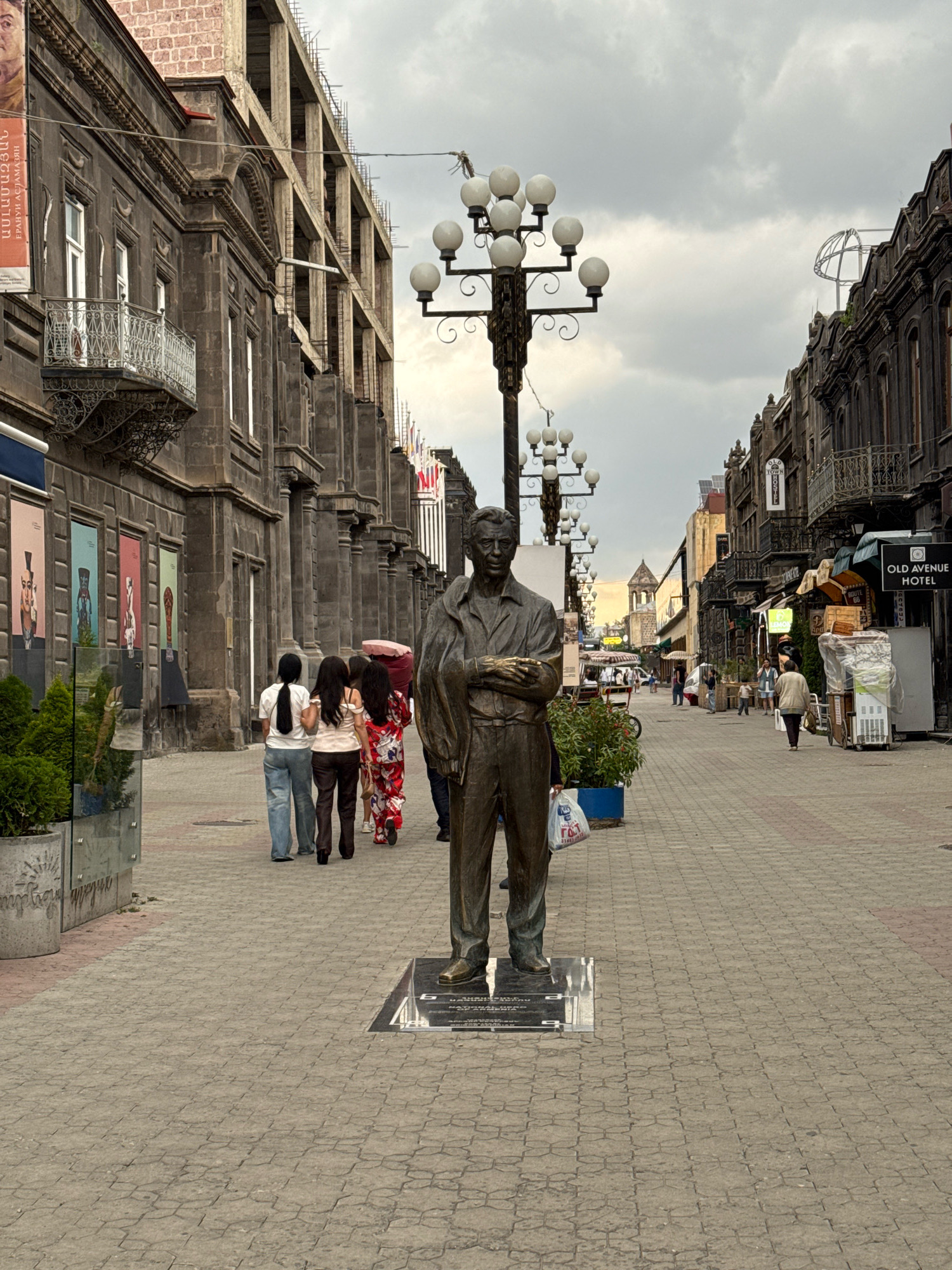 The image depicts a view down a pedestrian street in Gyumri, Armenia. In the foreground, a bronze statue of a man stands on a plinth. The man in the statue is depicted wearing a suit and holding an object in his hands. The street is paved with interlocking bricks and lined with buildings on either side.
The buildings on the left are older, dark-stone structures with ornate balconies and architectural details. Some of these buildings appear to be under renovation, with scaffolding and exposed brickwork visible. There are also posters and artwork displayed on the walls of some buildings.
The buildings on the right are also older, with similar architectural styles. A sign for "OLD AVENUE HOTEL" is visible on one of these buildings. Various businesses, including shops and what appear to be food vendors, line this side of the street.
Several people are walking along the street. On the left, three women are walking away from the camera, two of whom are in jeans and one in a patterned dress. Another group of people is visible further down the street.
There are lampposts with multiple globe lights lining the street, contributing to the urban atmosphere. The sky is overcast with grey clouds. The overall scene suggests a public space in a city, with a mix of historical architecture and ongoing development.