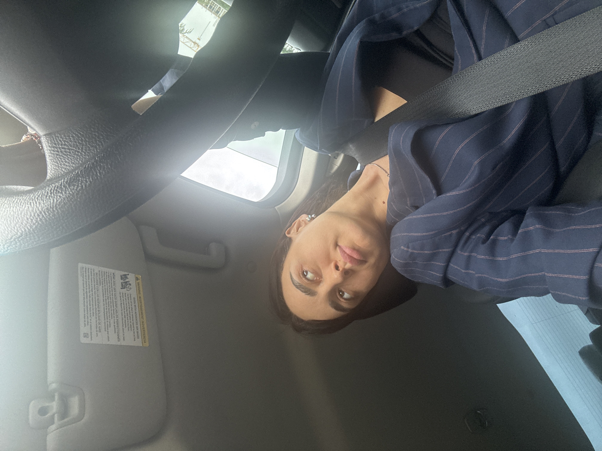 The interior of a car, likely a selfie taken by the driver. A person with long, dark hair and fair skin is visible wearing a navy blue pinstripe blazer and a seatbelt. A steering wheel and part of the dashboard are visible. A sun visor with warning labels is present. The location is unknown and cannot be determined from the image.