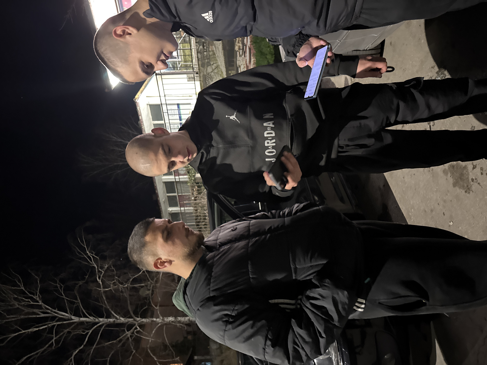 Three men are standing outdoors at night. The first man, located in the lower portion of the frame, has short, dark hair and a beard. He is wearing a black, puffy jacket with white stripes on the arm. The second man, located in the middle portion of the frame, has a shaved head and is wearing a black Jordan-branded sweatshirt and black pants. The third man, located in the upper portion of the frame, also has a shaved head and is wearing an Adidas-branded jacket and dark pants. The second man is holding a phone in his hand. The men are standing on a paved surface, possibly a sidewalk. In the background, there are bare tree branches and a building. The location is Topoli, Bulgaria.