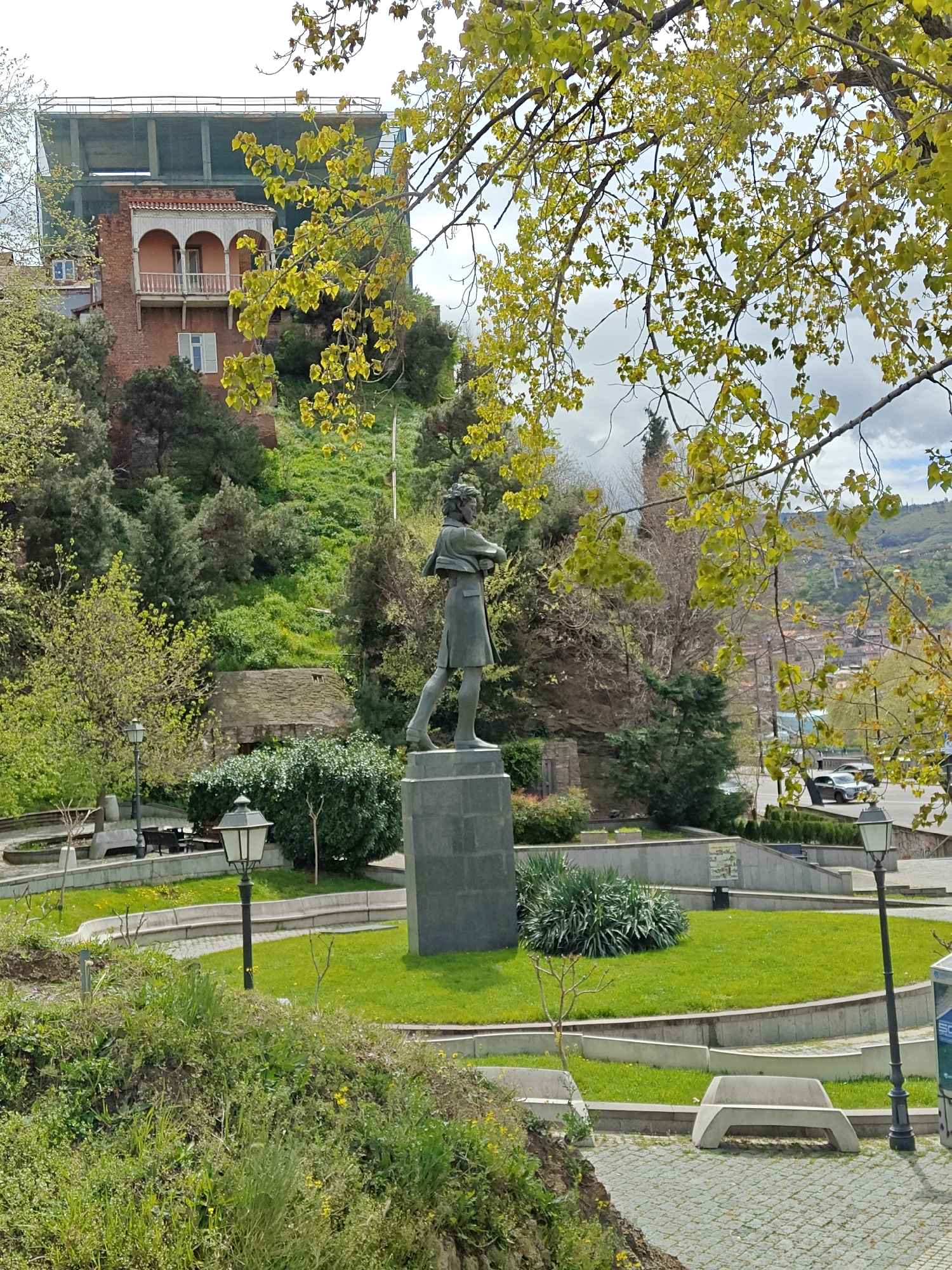 This image captures a scenic park or garden area in Tbilisi, Georgia, under bright daylight with a partly cloudy sky. The foreground is dominated by vibrant green foliage, including tree branches with fresh, light green leaves that frame the top and right of the frame, suggesting springtime.
A full-body statue, appearing to be cast in bronze, stands prominently on a dark, multi-tiered pedestal in the center. The figure, dressed in what appears to be a long coat or garment, is depicted in a thoughtful or observant pose, looking towards the right.
The park features well-maintained green lawns, paved walkways, and concrete benches. Several dark, traditional-style lampposts are visible throughout the space. Lush shrubs and trees are interspersed around the statue and along the pathways, contributing to the verdant atmosphere.
In the midground, a steep, green hillside rises, densely covered with trees and bushes. Nestled into this hillside is a multi-story brick building with a distinctive traditional wooden balcony featuring three arched openings, painted in a light pinkish-peach hue. Above this building, a large concrete structure is under construction, partially covered by green scaffolding netting.
The background reveals distant hills covered with greenery, and glimpses of a city or town with numerous buildings, hinting at the expansive urban landscape of Tbilisi. A road with parked cars is also visible in the distance on the right. No discernible human activity or legible text is visible in the frame.
