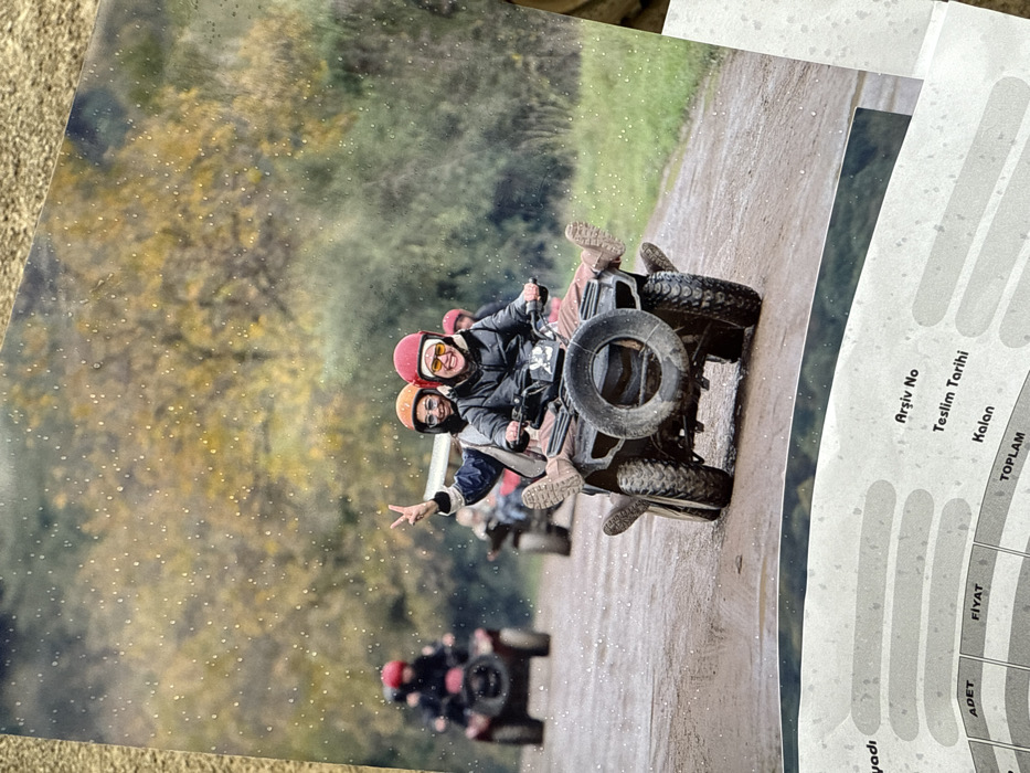 A printed photograph depicts a group of individuals riding all-terrain vehicles (ATVs) on a dirt road in Çiftlikköy, Turkey. In the foreground, two individuals are seated on a black ATV with a spare tire mounted on the rear. The person in the driver's position wears a red helmet, yellow goggles, and a black jacket, smiling and looking towards the viewer. The passenger, wearing an orange helmet, dark sunglasses, and a blue and white jacket, is smiling and making a peace sign with their left hand. Both have their legs extended and feet raised, wearing tan boots. The road surface is wet and muddy, with puddles visible. In the mid-ground, another ATV with two riders, one wearing a red helmet, is visible further down the road. Additional ATVs with riders are faintly visible in the background. The environment consists of dense autumn foliage with yellow and green trees lining both sides of the road. Fine white specks are visible across the image, potentially indicating rain or dust on the photograph itself. The lower portion of the image shows the top edge of a grey document with Turkish text, including "Arşiv No," "Teslim Tarihi," "Kalan," "ADET," "FİYAT," and "TOPLAM."