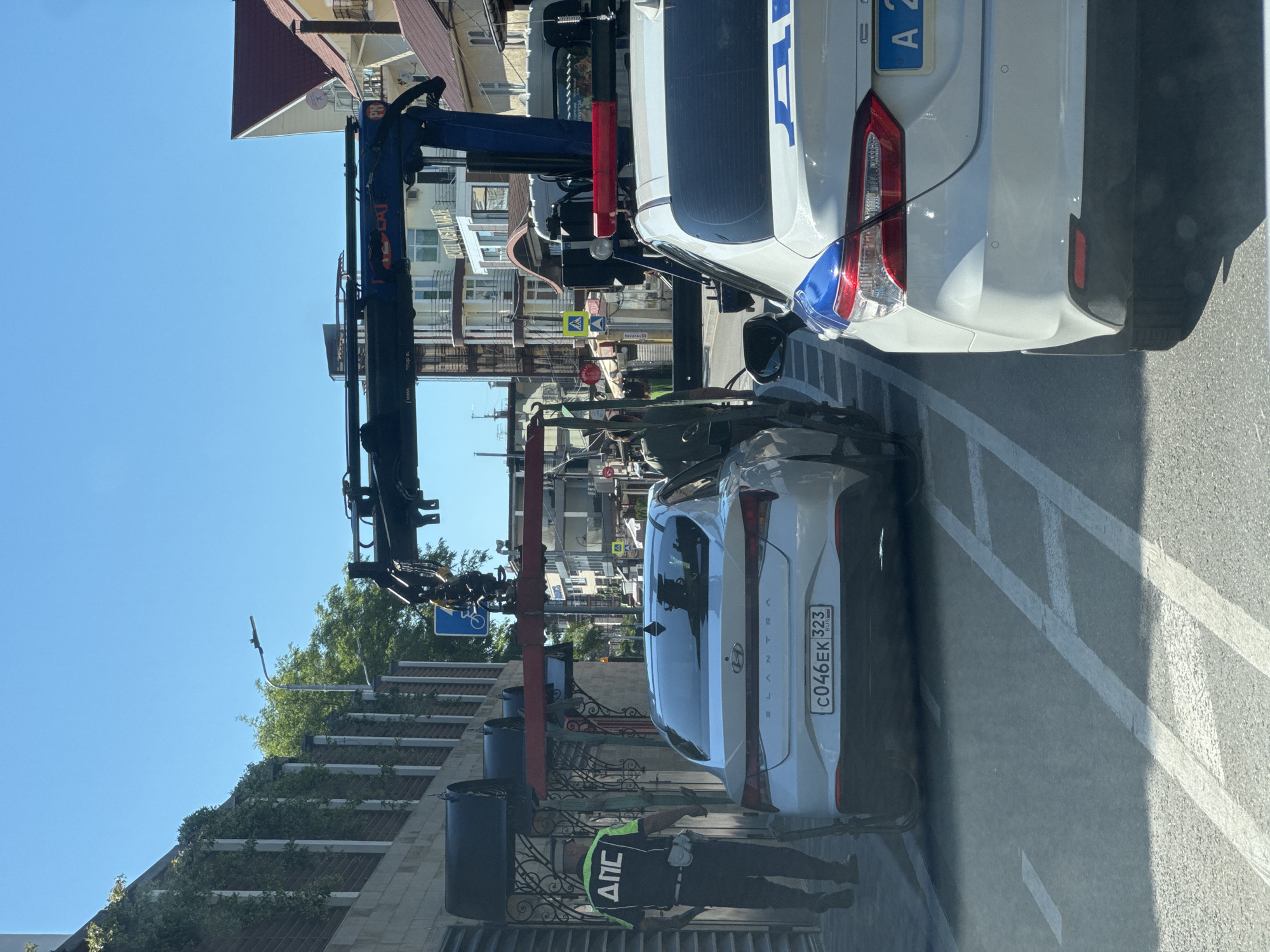 The image captures a street scene on a clear, sunny day, likely in Sochi, Russia. In the foreground, a white Hyundai Elantra sedan (license plate C046EK 323 RUS) is positioned in what appears to be a parking spot, marked by white lines on the asphalt. Behind it, a blue tow truck with a "Horyong" logo on its boom is preparing to tow the sedan.
To the right of the tow truck, the rear of a white van or service vehicle is visible, showing part of its license plate as "A 2". A male figure, dressed in a black uniform with a neon yellow vest bearing the Cyrillic text "ДПС" (DPS, indicating Russian Road Police or State Traffic Safety Inspectorate), stands near the sedan's rear, seemingly supervising the operation.
The background features several multi-story buildings under a bright blue sky, some with red-tiled roofs and balconies. Green foliage, possibly ivy, adorns parts of one building's facade. Street signs are visible, including a yellow triangular sign for a pedestrian crossing and a sign indicating "Эссовская 52" (Essovskaya 52). Another blue sign with a white bicycle symbol can also be seen. The bright sunlight casts prominent shadows on the ground and vehicles, suggesting a time between mid-morning and late afternoon.