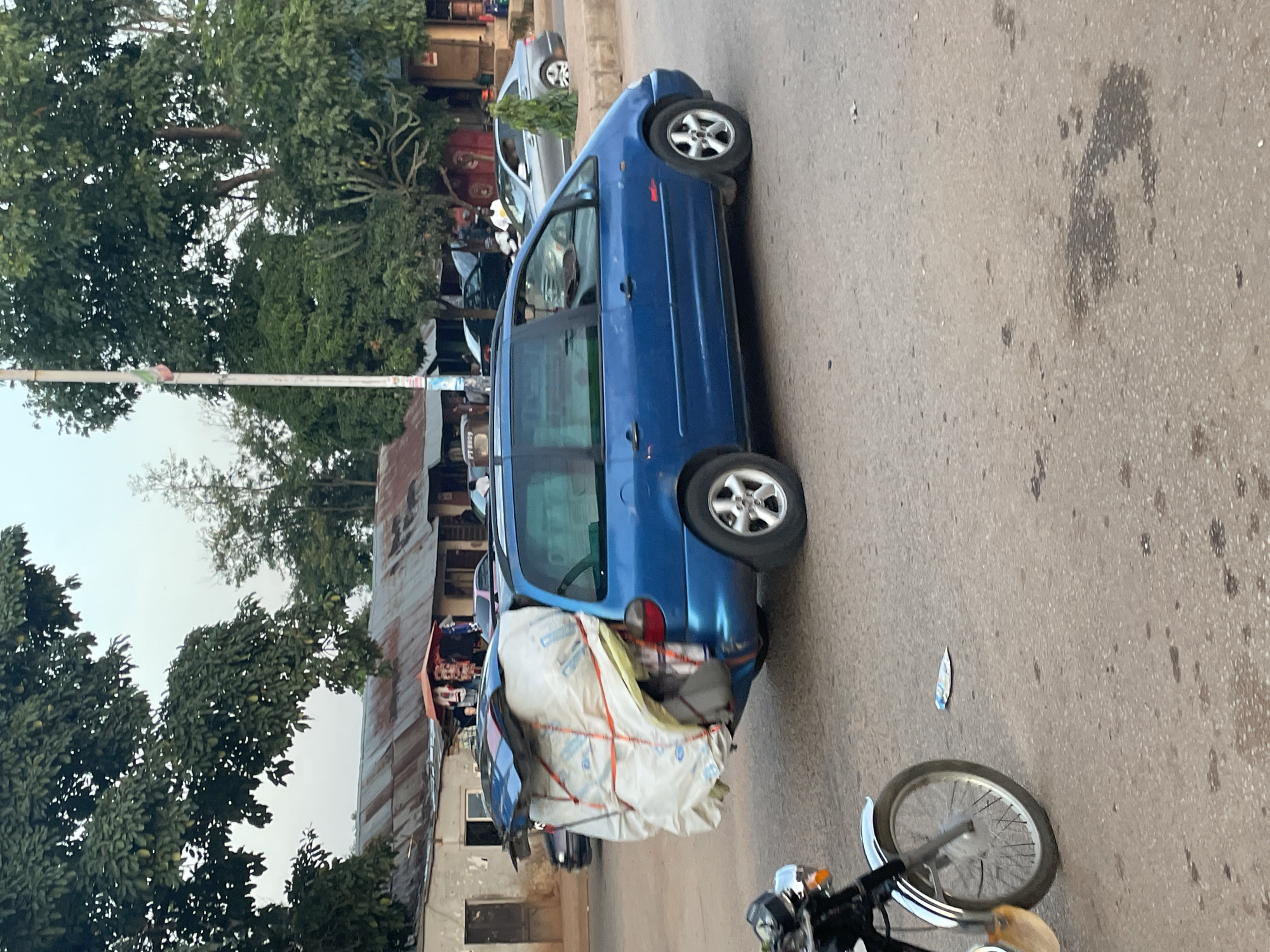 A blue minivan is parked on a paved road, its rear hatch open and overloaded with a large, white, tarp-wrapped bundle secured with orange straps. The bundle extends significantly beyond the back of the vehicle. A black motorcycle with chrome accents is partially visible in the foreground, parked to the left. In the background, there are buildings with corrugated metal roofs, trees, and other vehicles on the road. A few people are visible near the buildings. The sky is overcast. The scene appears to be in a commercial or residential area with some signs of activity.
