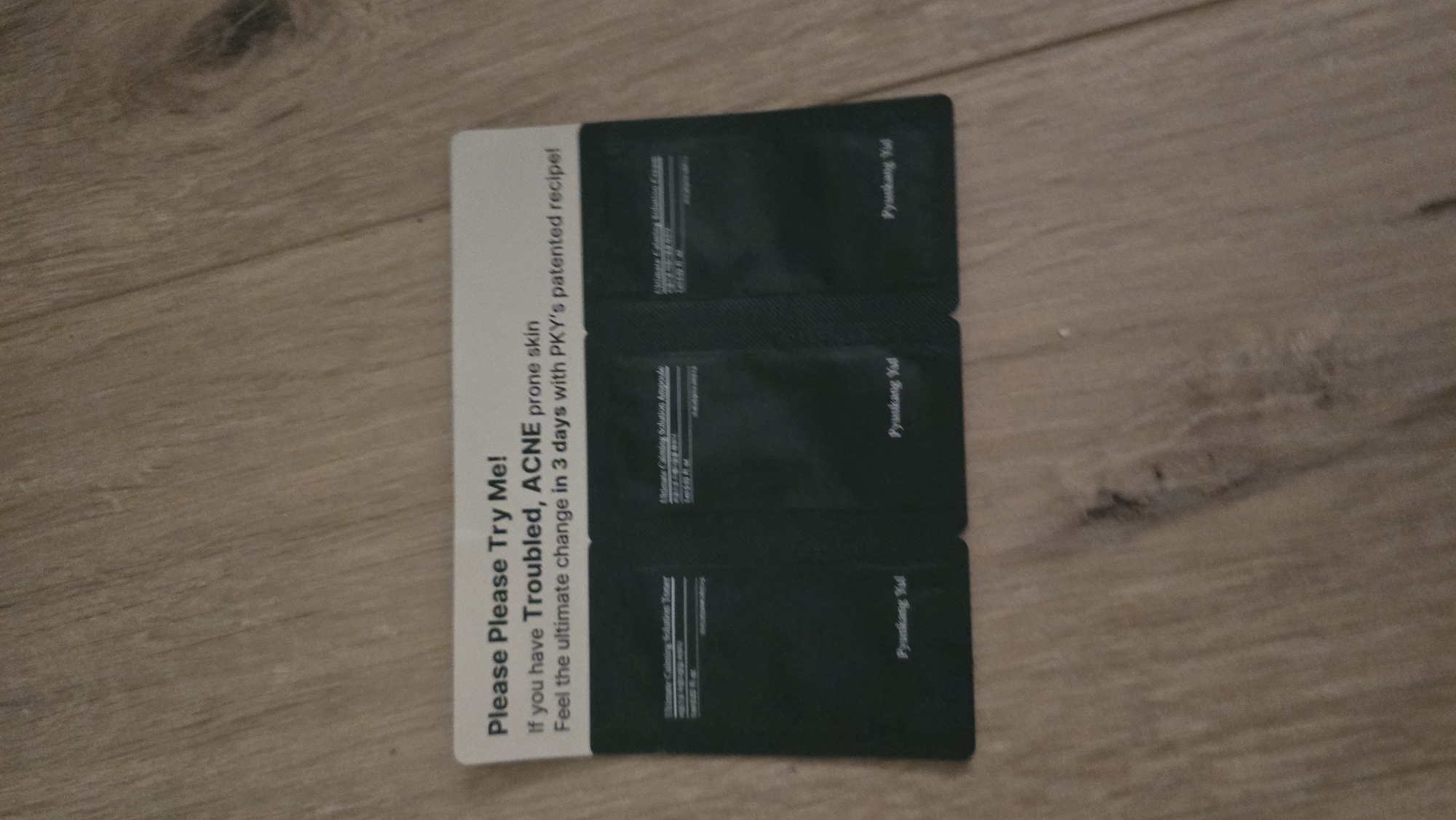 A small, dark card with white text is placed on a wooden floor. The card reads "Please Please Try Me! If you have Troubled, ACNE prone skin Feel the ultimate change in 3 days with PKY's patented recipe!". Below this text, there are three dark-colored sample packets. Each packet has white text printed on it, with the brand name "Pyunkang Yul" visible at the bottom of each. The wooden floor appears to be made of planks, and some debris is visible on the surface. There are no people or any other distinguishing features that would indicate a specific location in Batumi, Georgia.