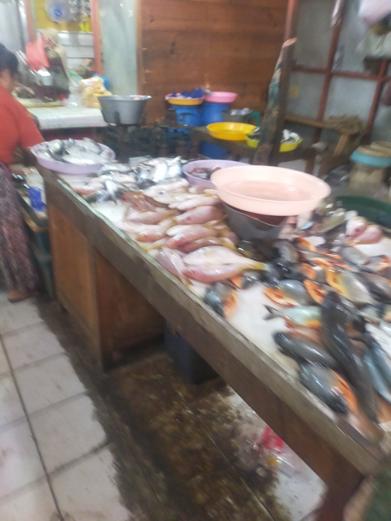 This is a blurred, medium shot of a fish market stall in Trayeman, Indonesia. The stall is a long wooden counter displaying a variety of fish on ice. There are pink and grey fish in the foreground, with some darker fish further back. A large pink bowl is on top of a grey scale at the back of the stall. To the left, a person wearing an orange shirt and a patterned skirt is partially visible, facing away from the camera. Behind the stall, there are wooden shelves with various items, including blue and yellow containers. The floor is tiled and appears wet.