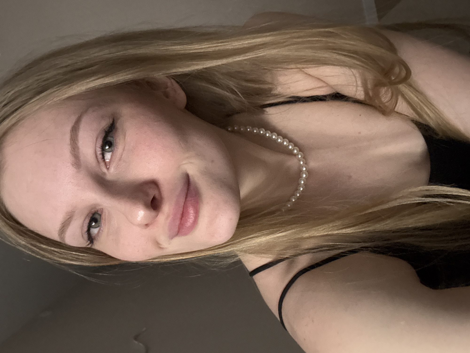 The image shows a close-up, slightly angled selfie of a young woman with blonde hair and blue-green eyes. She is wearing a black sleeveless top and a pearl necklace. Her lips are slightly parted and she has a subtle smile. Her hair is long and falls over her shoulders. The background appears to be an indoor setting with a neutral-colored wall. There is no discernible indication of the city of Perm, Russia.