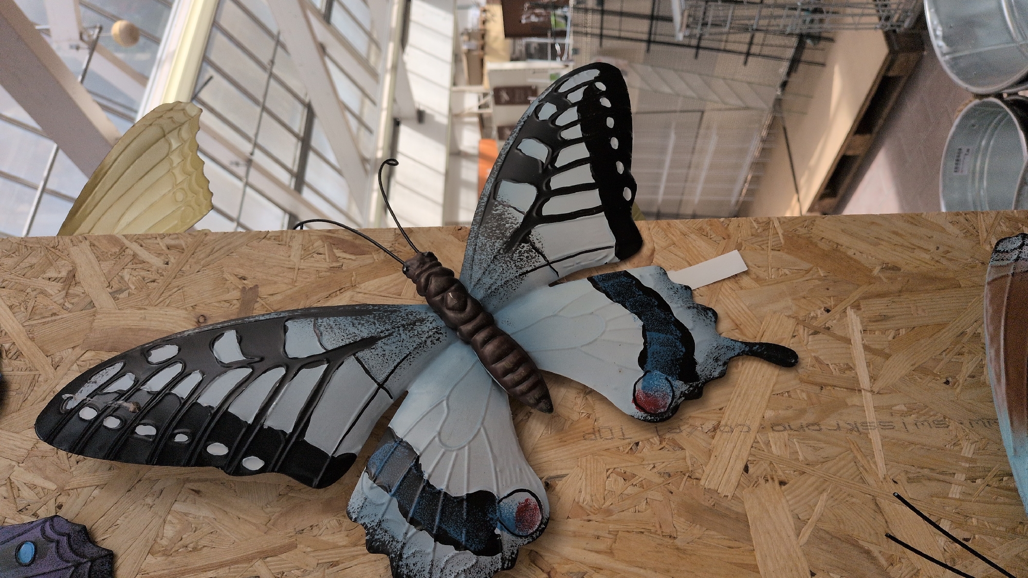 The image shows a decorative butterfly sculpture mounted on a wooden panel. The butterfly has black, white, and blue wings with a segmented brown body. Part of the background shows a greenhouse or store interior with shelves, pallets, and metal bins. The butterfly is the primary subject, and the wooden panel appears to be part of a display. The image was taken in Sömmerda, Germany.