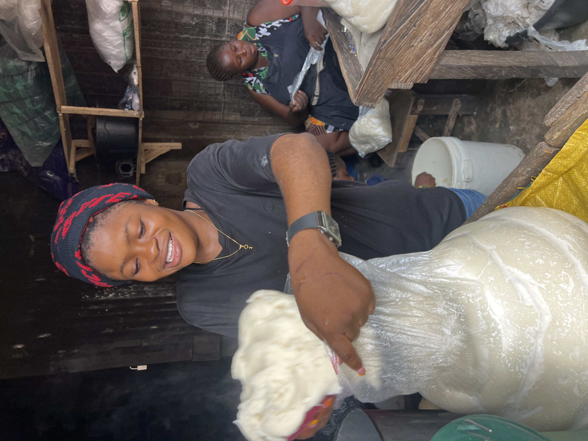 In Nkpor, Nigeria, a young woman with a black and red beanie smiles as she holds a large bag filled with a white, dough-like substance. She wears a black t-shirt and blue denim shorts and a silver watch on her left wrist. In the background, another woman sits and appears to be wrapping something in plastic. The scene takes place in what looks like an outdoor market stall, with shelves stocked with various items, including bags of produce and plastic containers. A wooden counter with a yellow cloth is visible in the foreground. The ground is dirty, suggesting a typical market environment. The overall atmosphere is one of daily commerce and food preparation.