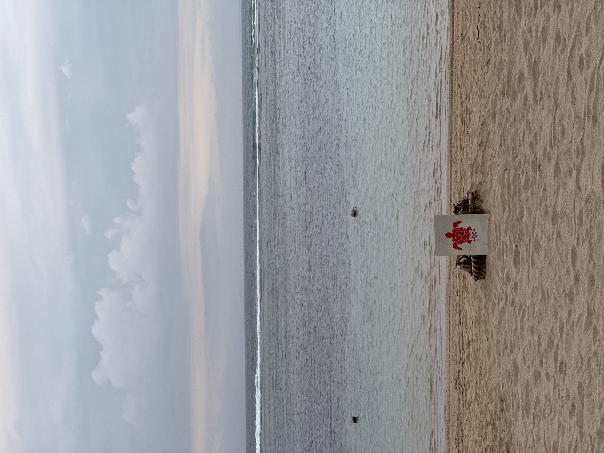 The image depicts a serene beach scene in Kuta Selatan, Indonesia, under an overcast sky. The foreground is dominated by the textured sand of the beach, leading to the calm, greyish-blue water of the ocean. A thin line of white surf breaks on the horizon, separating the water from the sky. In the middle ground, a sign with a red turtle emblem is placed on the sand, propped up by what appears to be woven material. Two buoys are visible in the water, one closer to the shore and another further out. The overall atmosphere is peaceful and slightly subdued due to the cloudy weather.