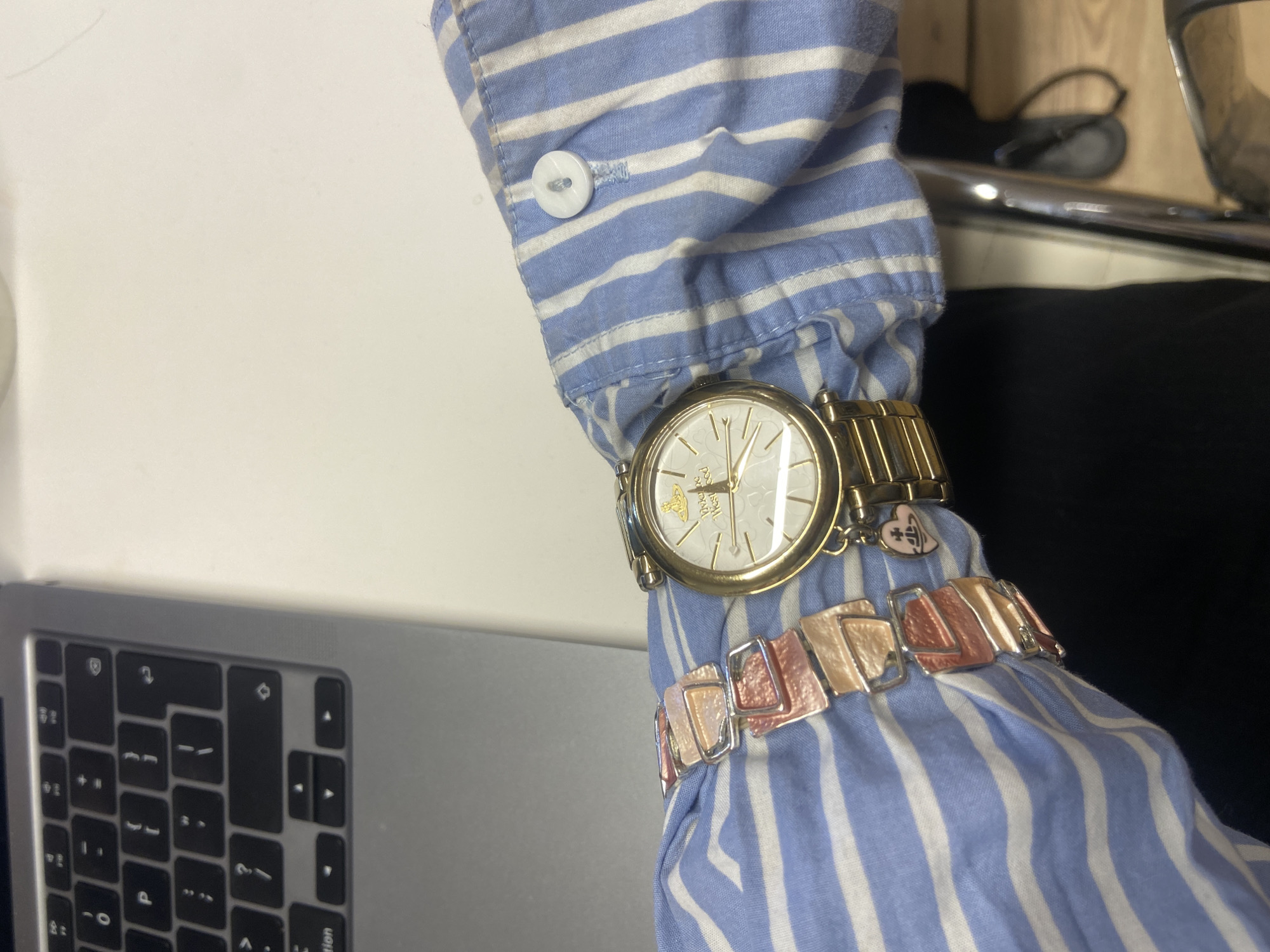 A close-up shot shows a person's arm wearing a striped blue and white shirt, adorned with a gold wristwatch and a silver and gold bracelet. The watch is prominently featured, with a gold band and a white face displaying the brand "Vivienne Westwood." A silver and gold bracelet is worn next to the watch. The arm is positioned near a laptop keyboard, with the left side of the keyboard visible. The setting appears to be indoors, possibly an office or home workspace. The overall lighting is bright and even.