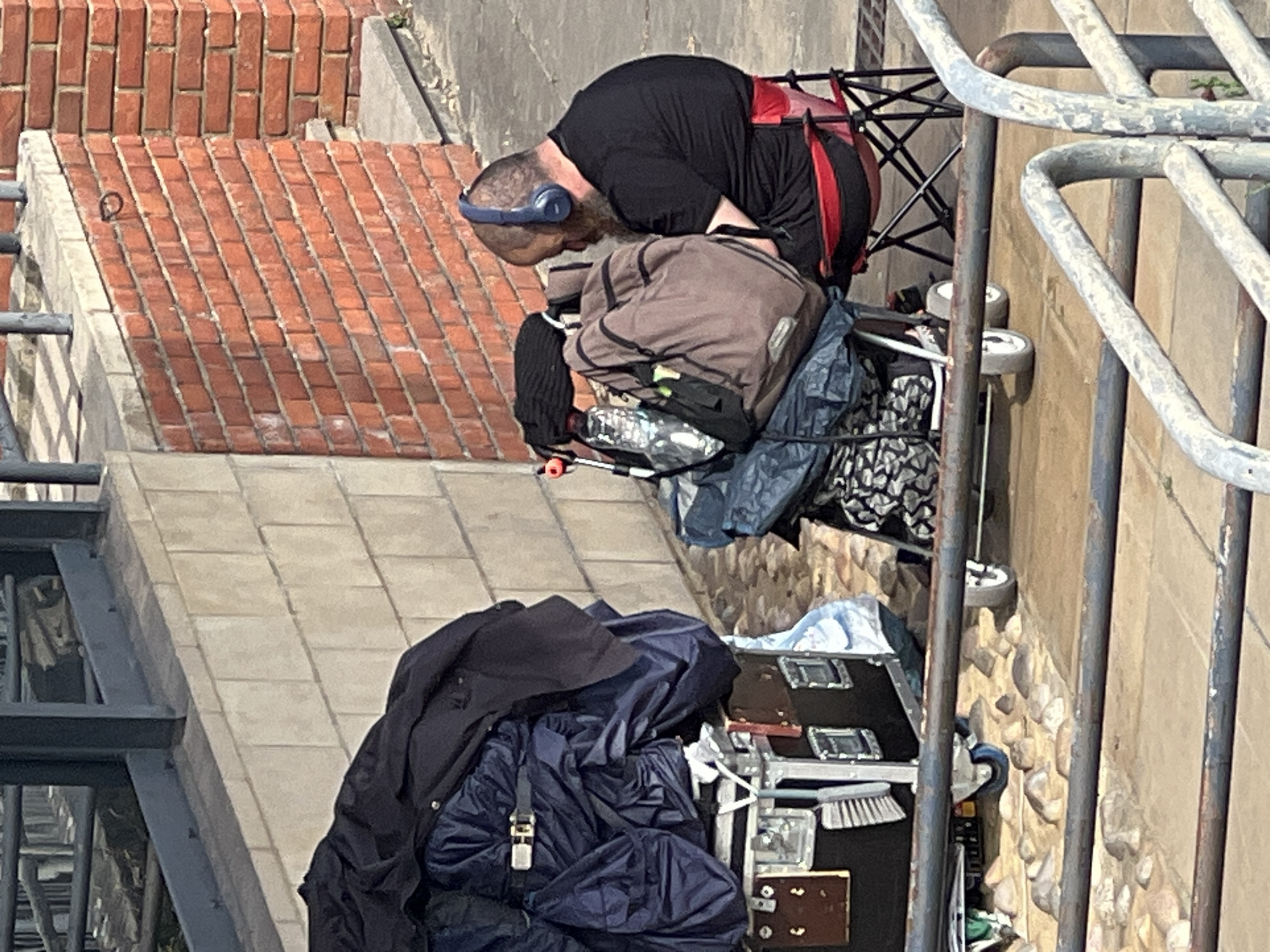 An outdoor, elevated view shows a man sitting on a red folding chair next to a cart piled with belongings. The man has a beard and is wearing headphones and a black t-shirt. He is looking down and to his left. The cart contains bags, a water bottle, and other items. To the left of the cart is a large black case with wheels, also covered with dark blue fabric. The background features a brick wall and a lighter colored stone wall. The scene appears to be in a public area. The image was taken in Guildford, United Kingdom.