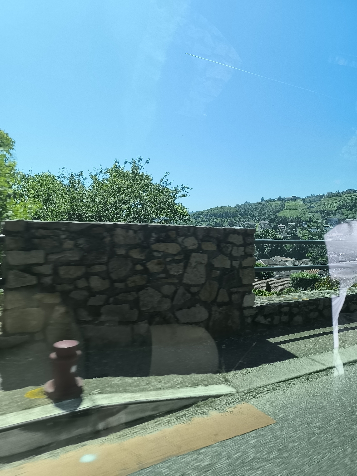 The photograph, seemingly taken from a moving vehicle, presents a partial view of Chavanay, France. The composition is divided into three primary sections: foreground, middle ground, and background.
Foreground: A stone retaining wall dominates the lower portion of the image, with a small, red, cylindrical object placed on the adjacent ledge. A significant portion of the lower frame consists of the road surface, a blurred indication of the vehicle's movement.
Middle Ground: Behind the retaining wall, a section of wrought-iron fencing is visible. A cluster of trees and shrubs provides a backdrop, partially obscuring the landscape beyond.
Background: Rolling hills characterize the horizon, dotted with houses and what appears to be terraced vineyards, typical of the Rhône-Alpes region where Chavanay is located. The sky is a clear, bright blue, with a contrail indicating aircraft activity.
Reflections are present in the image.