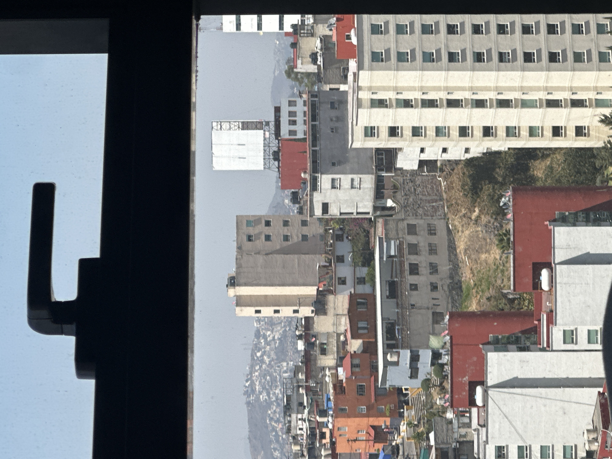 The image depicts an urban landscape in Huixquilucan de Degollado, Mexico, viewed through a window frame with a dark handle visible at the top. The scene is dominated by a dense concentration of buildings spanning varied architectural styles and heights, conforming to a hilly topography under a hazy, light blue sky.
In the foreground and mid-ground, numerous multi-story structures are visible. These include residential and potentially commercial buildings rendered in shades of beige, gray, white, orange, and red. Many rooftops feature multiple white or blue cylindrical water storage tanks (tinacos) and several white horizontal gas tanks.
Prominently, a tall, slender beige building with multiple square windows is situated on the mid-right. Further into the mid-ground, a large, blank white billboard, supported by a metal truss structure, stands atop a red-roofed building. Another tall, lighter-colored building with repetitive window patterns is partially visible on the far right.
The urban development extends up sloping terrain, with areas of sparse, dry vegetation and exposed earth visible between structures. In the distant background, a large mountain range is densely covered with numerous smaller, tightly packed residential structures, indicating extensive urban sprawl. No individuals or discernible activities are depicted. The overall impression is that of a sprawling, established city during daylight hours.