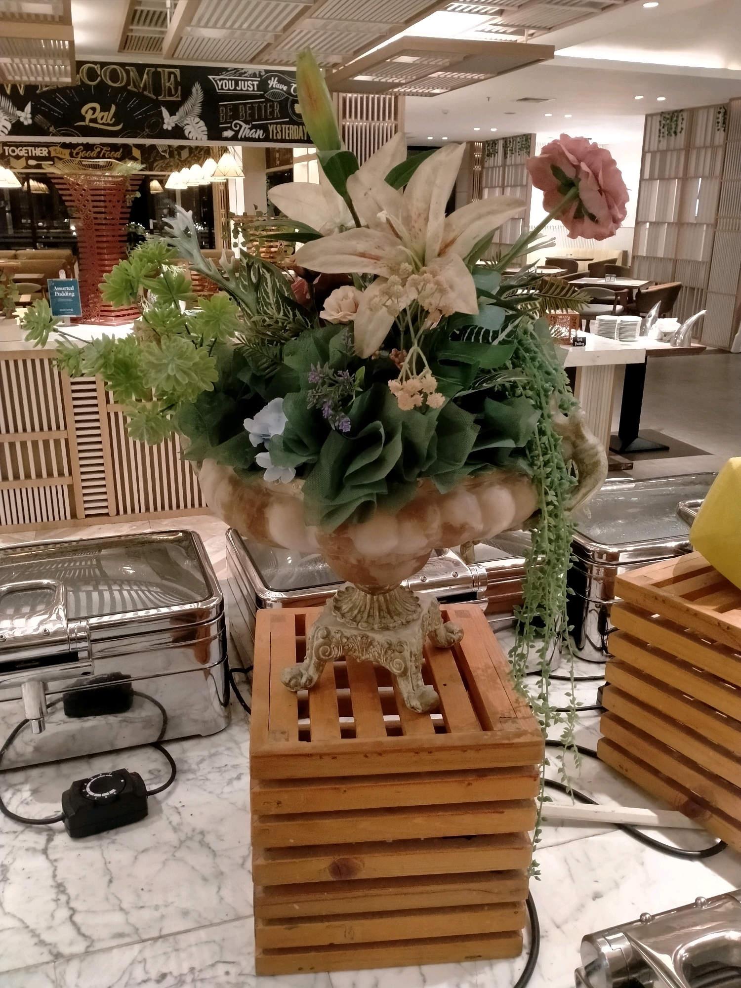 The image shows a buffet spread in a restaurant, likely in Batu, Indonesia, given the context. The foreground is dominated by a large floral arrangement in an ornate vase, placed on a wooden stand. To the left, there are two stainless steel chafing dishes with glass lids, one of which has a black temperature control device attached. To the right, there are stacks of wooden crates. In the background, a sign with the words "WELCOME Pal" and "YOU JUST Have BE BETTER Than YESTERDAY" is visible, along with dining tables and chairs. The overall setting suggests a dining establishment.