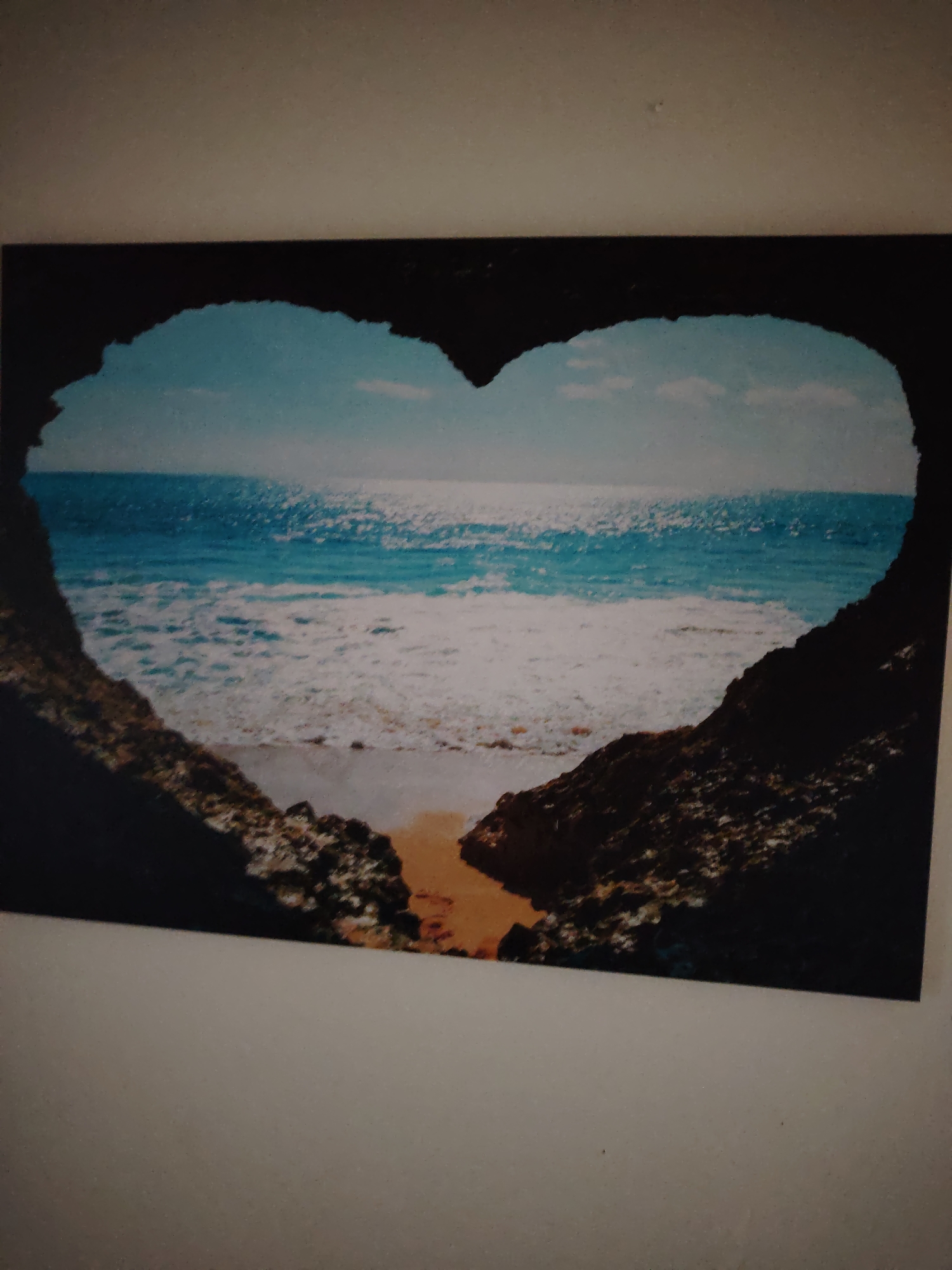 The image is a framed photograph hanging on a plain, light-colored wall. The photograph depicts a seascape framed by dark, rocky formations that create a heart shape. Through this heart-shaped opening, a bright blue ocean meets a sandy beach. The ocean has white-capped waves and reflects the sunlight, creating a shimmering effect. The sky above is a lighter blue with a few wispy clouds. The framing of the image suggests it might be a window or an opening in a cave looking out onto the sea. There are no people visible in the image. The scene is serene and natural, evoking a sense of peace and beauty. There is no information to suggest the location is in the city of Huntingdonshire, United Kingdom.