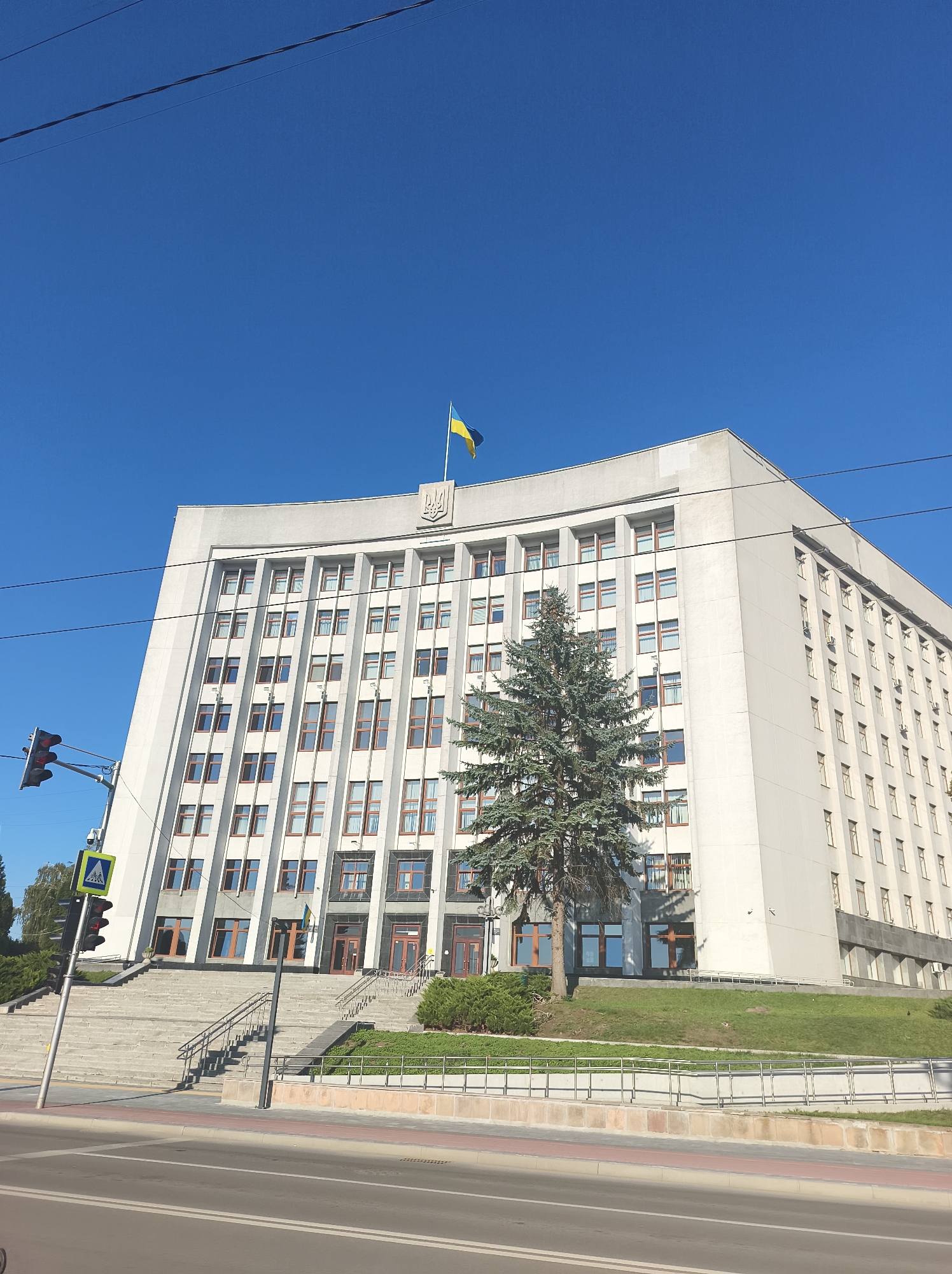 The image shows the Ternopil Regional State Administration building in Ternopil, Ukraine, under a clear blue sky. The building is a large, modern, concrete structure with multiple stories and numerous windows. A Ukrainian flag is prominently displayed on the rooftop, along with the Ukrainian coat of arms. In front of the building, there is a wide staircase leading up to the entrance, flanked by greenery. A large evergreen tree stands to the left of the staircase, partially obscuring the building. A traffic light and a pedestrian crossing sign are visible on the left side of the image, indicating a street in front of the building. The street itself, with road markings and a sidewalk, occupies the foreground of the image. Overhead power lines are also visible against the sky. There are no people visible in the image.