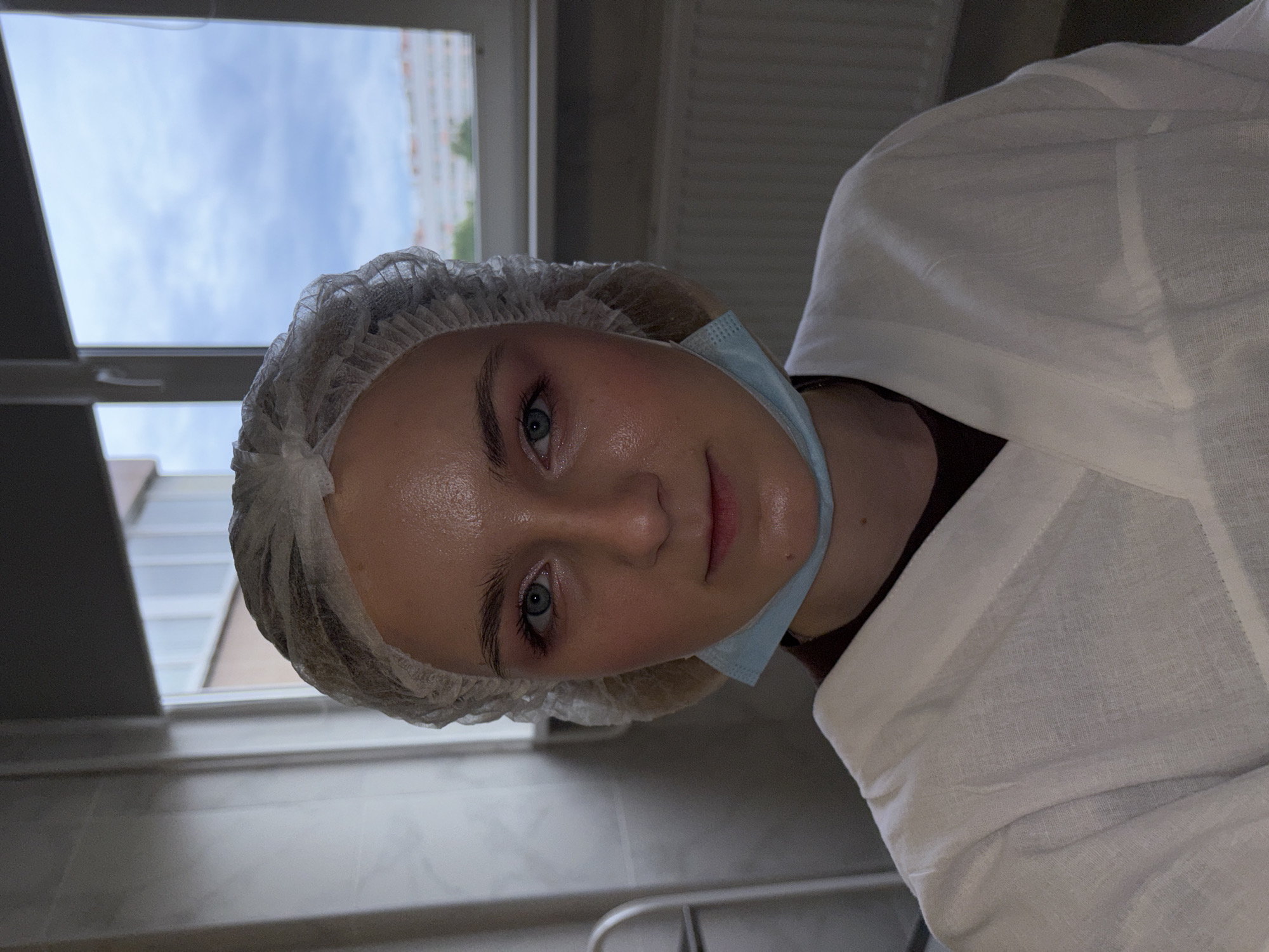 A portrait-oriented image shows a young woman looking directly at the camera. She has fair skin, light blue eyes, and light makeup, including mascara. Her hair is covered by a white, disposable bouffant cap, and a light blue surgical mask is pulled down, resting under her chin. She is wearing a white, possibly linen, smock or gown, over a darker garment.
The setting appears to be indoors, likely a medical, clinical, or beauty treatment environment. A large window in the upper left background reveals a cloudy sky and distant buildings, indicating it is daytime. The interior background is plain, with some indistinct fixtures visible in the lower right. The woman's expression is calm and neutral. No text is visible in the image.