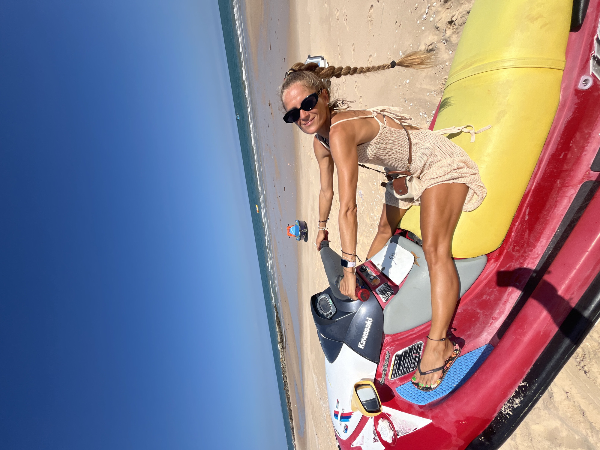 A woman with blonde hair in a braid, wearing sunglasses and a beige knitted dress, sits on a red and yellow jet ski on a sandy beach. Her left leg is extended, and her right leg is bent at the knee. She is holding the handlebars of the jet ski and looking towards the camera with a smile. The ocean and a clear blue sky are visible in the background. The location is identified as Phu Thuy Ward, Vietnam.