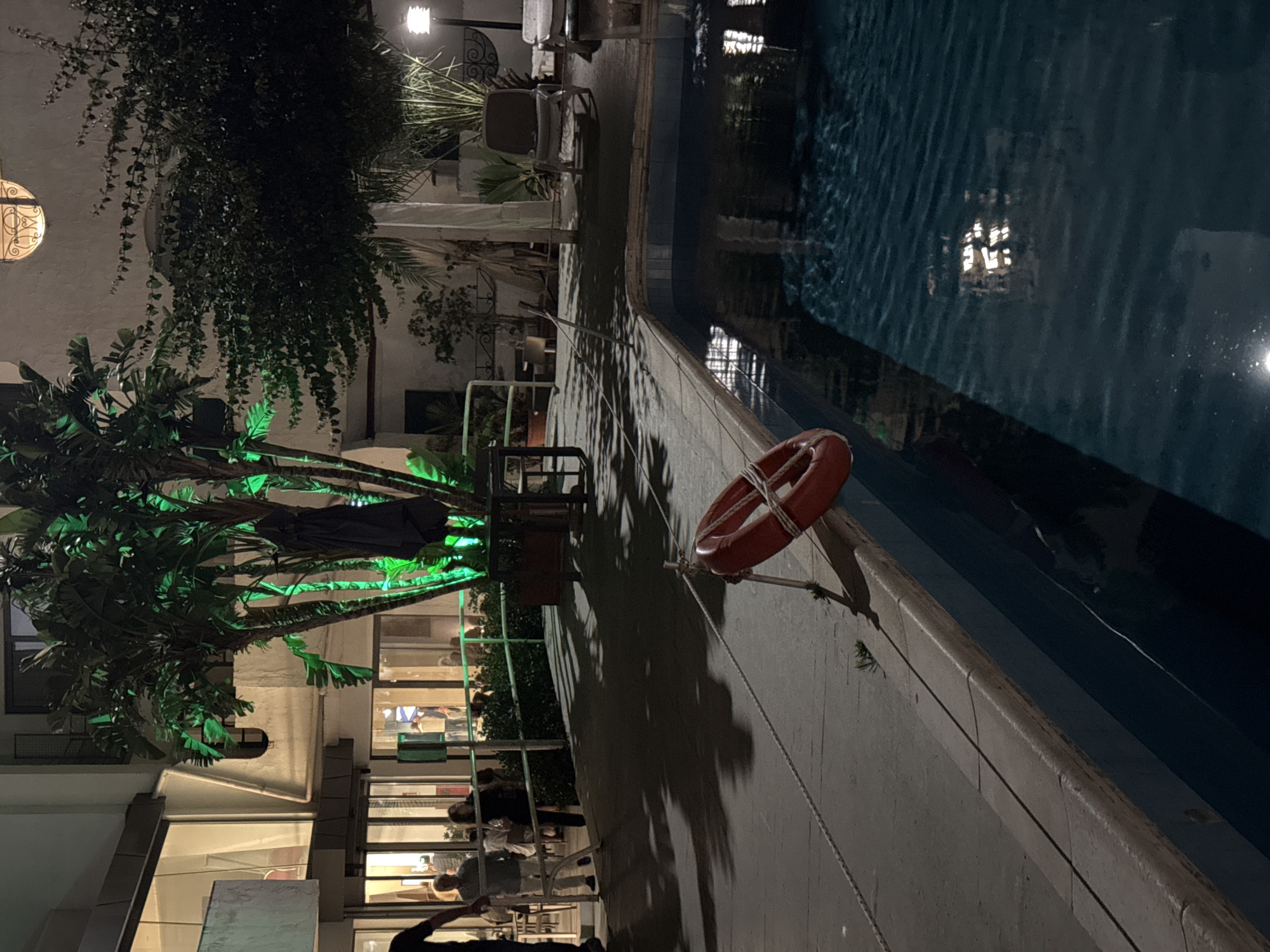 The scene depicts an outdoor evening view in Isola delle Femmine, Italy, likely a hotel or resort courtyard with a swimming pool. In the lower right, a rectangular swimming pool with dark blue water reflects internal building lights. A red lifebuoy is mounted on a pole leaning over the pool's edge in the lower-left foreground, with a white rope extending horizontally across the dark paved pool deck.
The midground features a paved walkway displaying distinct shadows from overhead foliage. A metal railing delineates this walkway from a lower area adjacent to a multi-story building. Several individuals are present: a partial view of a person in a dark shirt on the far left, an older male in a light shirt facing right, and a female in dark attire facing left. Multiple indistinct figures are visible inside the building through large, illuminated windows.
A prominent multi-trunk tree with dense foliage, illuminated by green lights, stands centrally, with a dark, folded umbrella positioned beside its base. To the right of the pool, two light-colored lounge chairs and a modern lamppost emitting white light are situated amidst various potted plants and dense foliage. The background consists of the multi-story building with numerous illuminated windows, and an upper section features an ornate, semicircular window with a decorative grate.