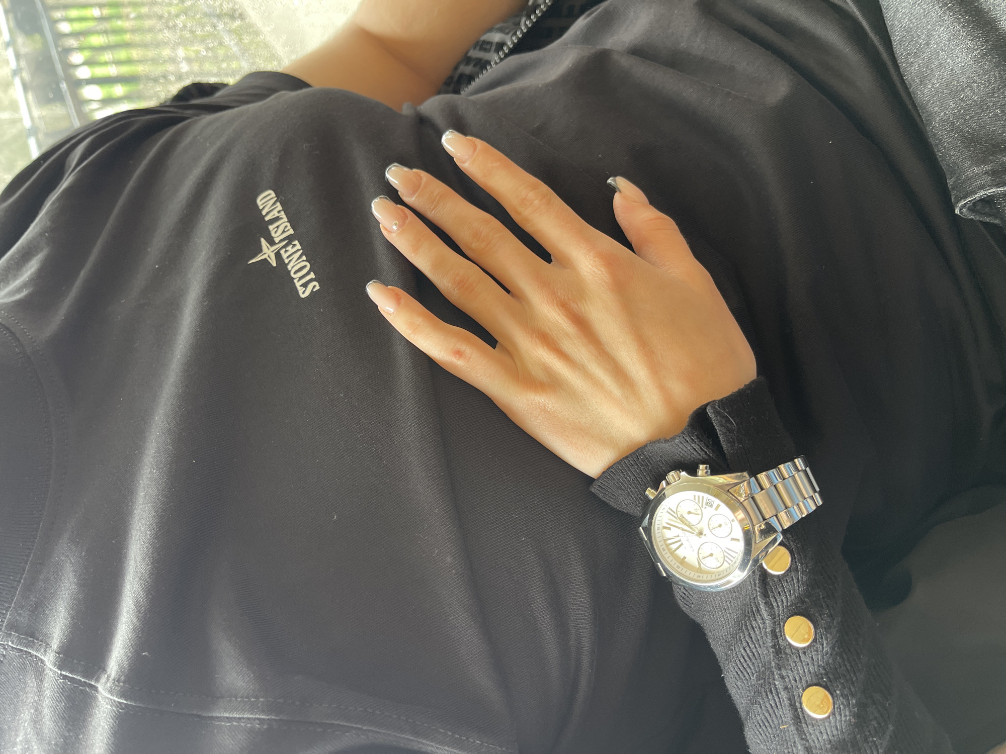 A close-up shot reveals a person reclining, clad in a black Stone Island shirt. The individual's right hand rests on the shirt, adorned with neatly manicured nails. A silver wristwatch with a metal band is visible on the wrist of the person wearing a black sweater with 4 gold buttons. The scene appears to be indoors with a glimpse of greenery visible outside the window. The location in the city of Veliko Tarnovo, Bulgaria is not discernible from the image.
