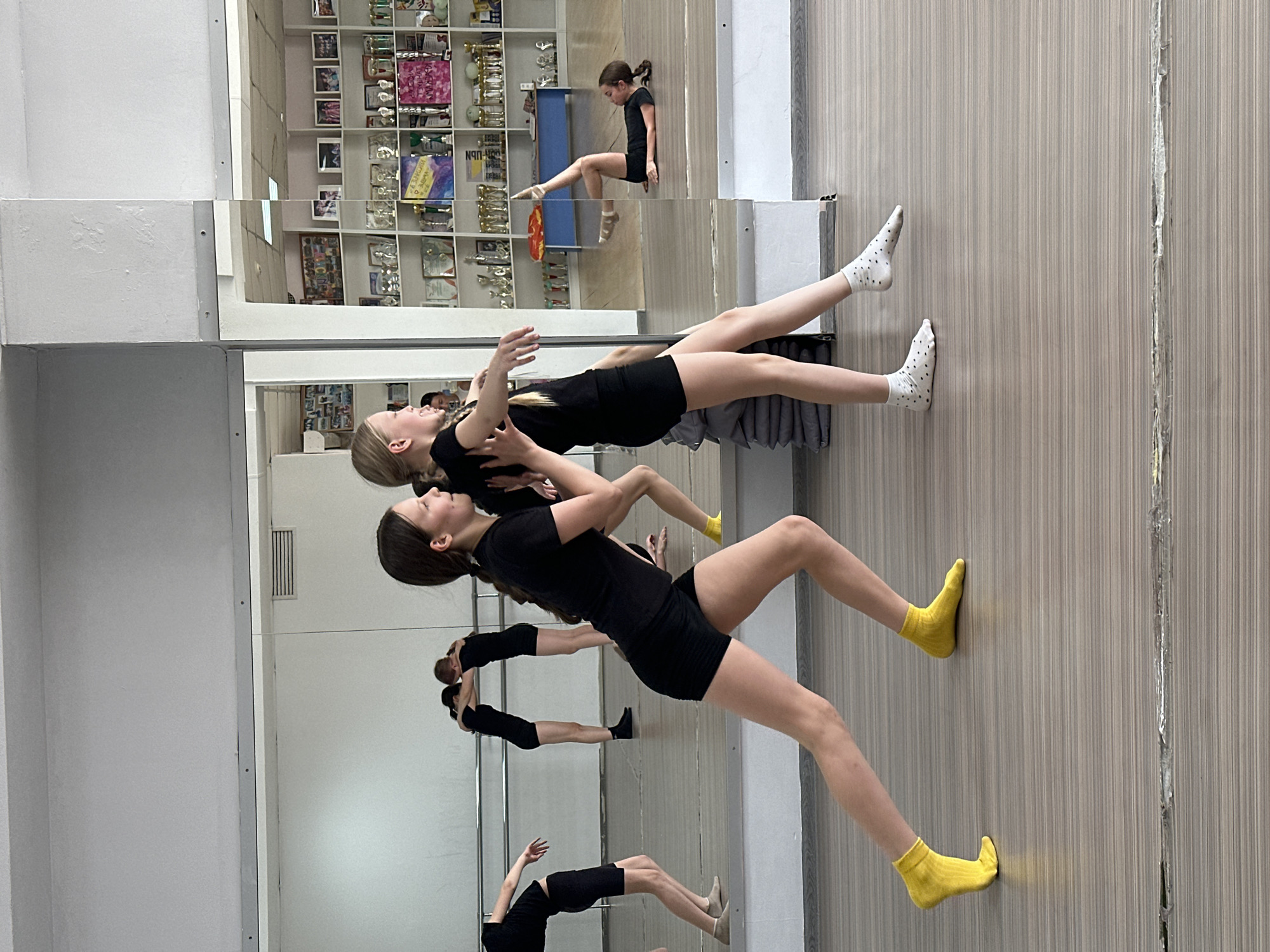 The image captures a dance studio scene. Two young girls, dressed in black leotards and shorts, are engaged in a dance exercise. The girl at the bottom is wearing bright yellow socks, while the girl at the top is wearing white socks with black dots. They are positioned closely together, with the lower girl supporting the upper girl. The studio has mirrors on the walls, reflecting their movements and creating multiple images of the dancers. In the background, there's a wall with shelves displaying trophies and framed photos. The floor is a light-colored wood laminate. The image was taken in a studio located in Votkinsk, Russia.