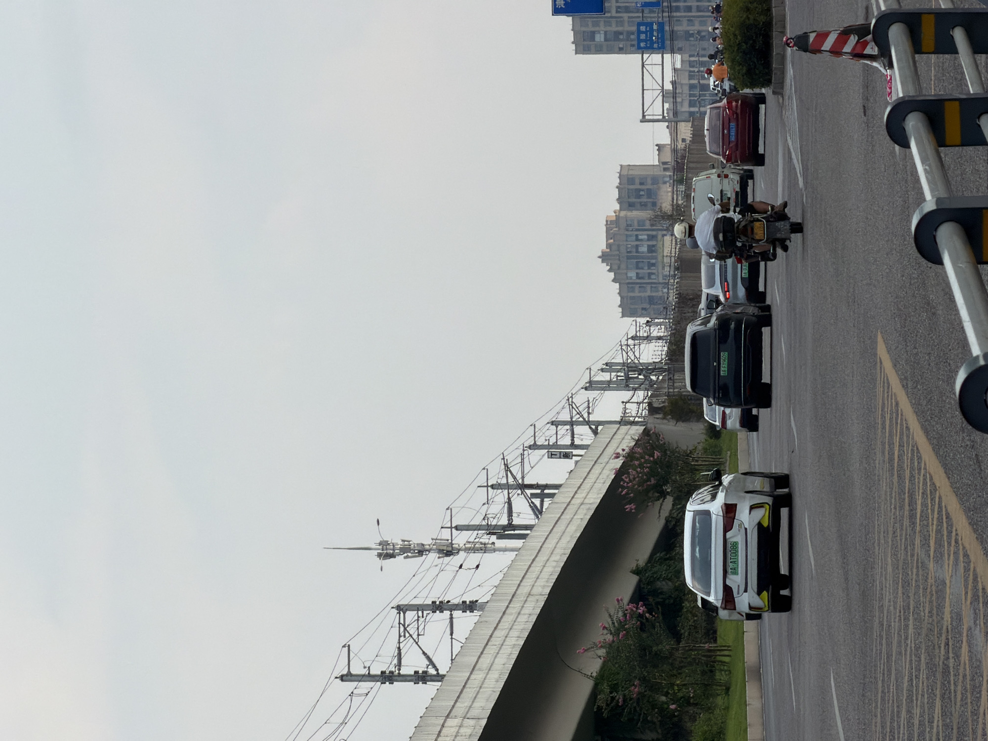 In Jiangjin, China, a road scene unfolds with multiple vehicles in transit under an overcast sky. The foreground shows a traffic lane with yellow and white markings. A white sedan with a license plate ending in "0086" is moving away from the camera. To its right, a dark-colored SUV is also in motion. A motorcycle with a rider wearing a white helmet and dark shorts is in the middle lane, also moving away. Behind the motorcycle, a white van and a red sedan are visible. In the background, elevated train tracks supported by concrete structures run above a street lined with greenery and flowering bushes. Further back, urban buildings of varying architectural styles rise against the hazy sky. A blue traffic sign with Chinese characters is present on the right side of the road.