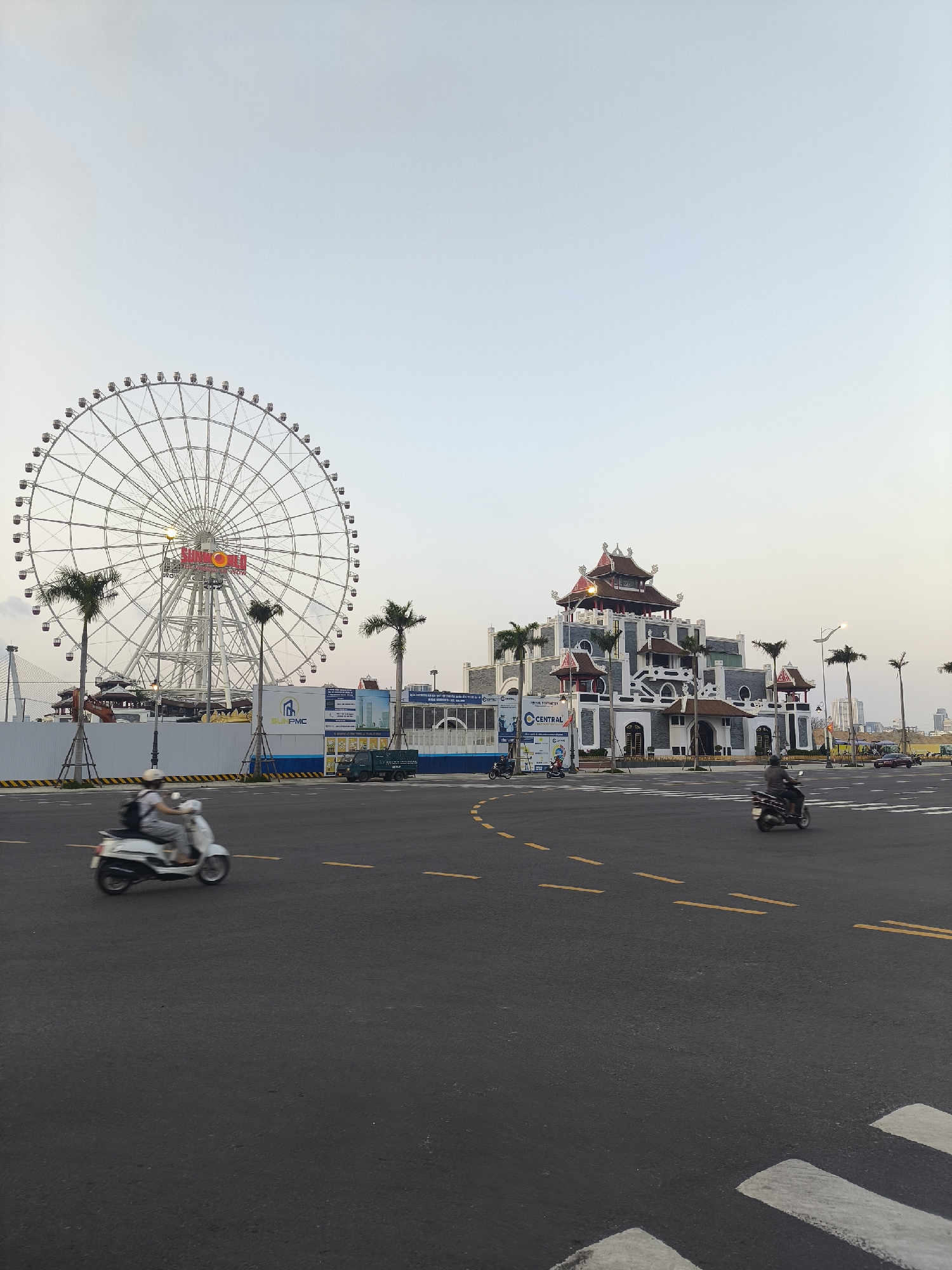 The image displays an urban scene in Đà Nẵng, Vietnam, focusing on the Sun World Danang Wonders (Asia Park) complex. On the left, the prominent Sun Wheel Ferris wheel, a large white structure with red "SUN WORLD" signage, dominates the skyline. To the right, a multi-tiered building with traditional Vietnamese architecture, featuring dark gray and white facades and red-tiled roofs, is visible. The foreground and midground consist of a wide, dark asphalt road with yellow dashed lane markings and white crosswalk stripes. Several motorbikes are in motion; a white scooter carrying two helmeted individuals, the passenger with a backpack, moves from left to right in the left foreground, exhibiting motion blur. Another dark scooter with a single rider is similarly blurred in the right midground, also moving left-to-right. Tall palm trees are strategically placed near both main structures. In the background, behind white corrugated construction barriers marked with "SUN PMC" and "CENTRAL" signage, an excavator and a dark green truck are partially visible. The sky is a clear, light blue, indicative of late afternoon conditions.