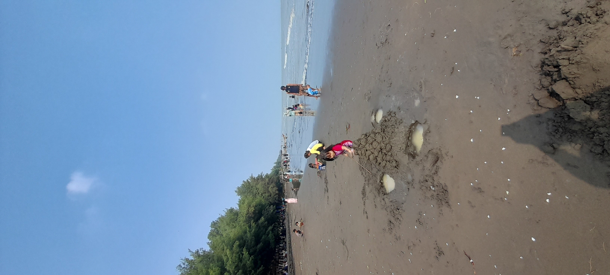 In Nyamplung Sari, Indonesia, a wide, sandy beach is populated with numerous people enjoying the day. In the foreground, a young girl in a red and pink striped dress is actively building a sandcastle. She is using a stick to shape the wet sand, which has created several small puddles around the base of the structure. Behind her, other children and adults are scattered across the beach. Some are playing in the shallow water near the shoreline, while others are closer to the tree line that borders the beach. The sea is calm with gentle waves, and the sky above is a clear blue with a single, small white cloud. The overall scene depicts a lively and relaxed atmosphere at a coastal recreational area.