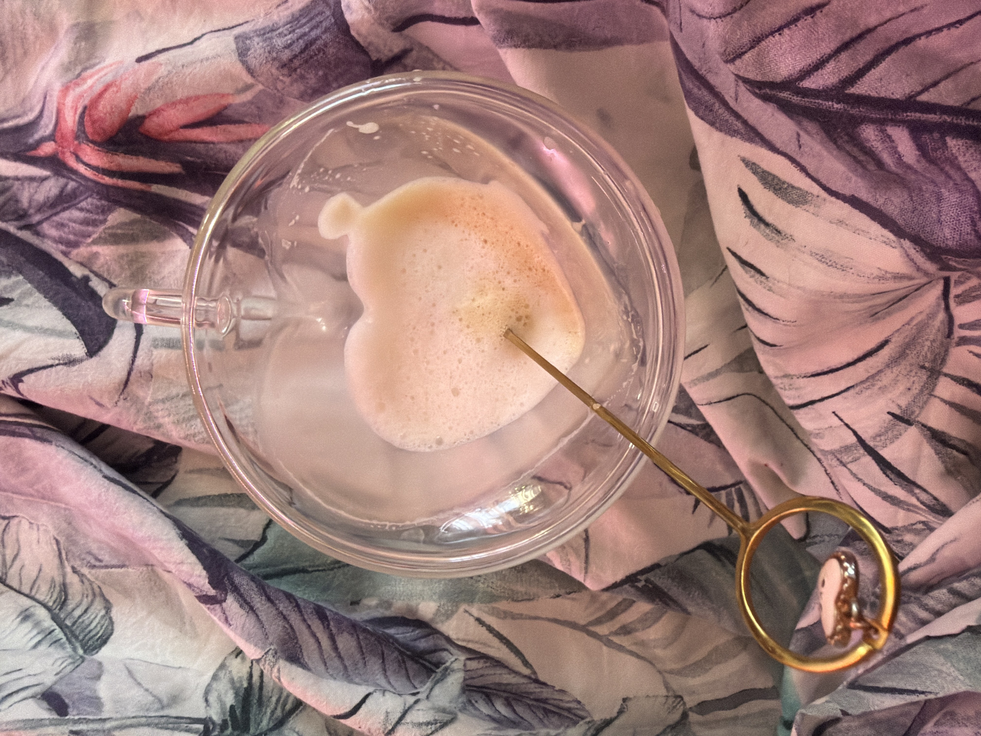 A top-down close-up shot reveals a clear glass mug containing a small amount of liquid and a dollop of foam shaped like a heart. A gold-colored stirring stick with a circular loop handle and a small pendant hangs over the mug. The mug and utensil rest on a patterned fabric with designs of leaves in shades of pink, gray, and white. The scene is lit with a soft, diffused light. No people are visible. The specific location within Chaikovsky, Russia, is not discernible from the image.
