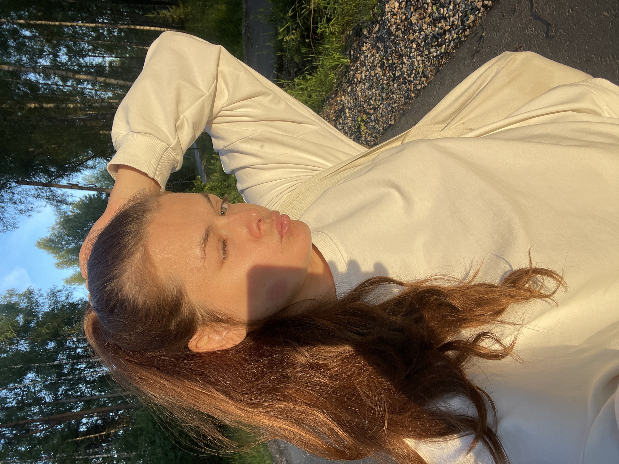 A young woman with brown hair in a ponytail is posing for a selfie outdoors. She is wearing a cream-colored sweatshirt. Her left hand is raised, touching her head. Her face is turned to the side, and she is winking and puckering her lips. There is a purplish mark on her left cheek. The background consists of trees and a clear sky. The lighting suggests it is either early morning or late afternoon. The location is stated to be in the city of Zarechny, Russia.