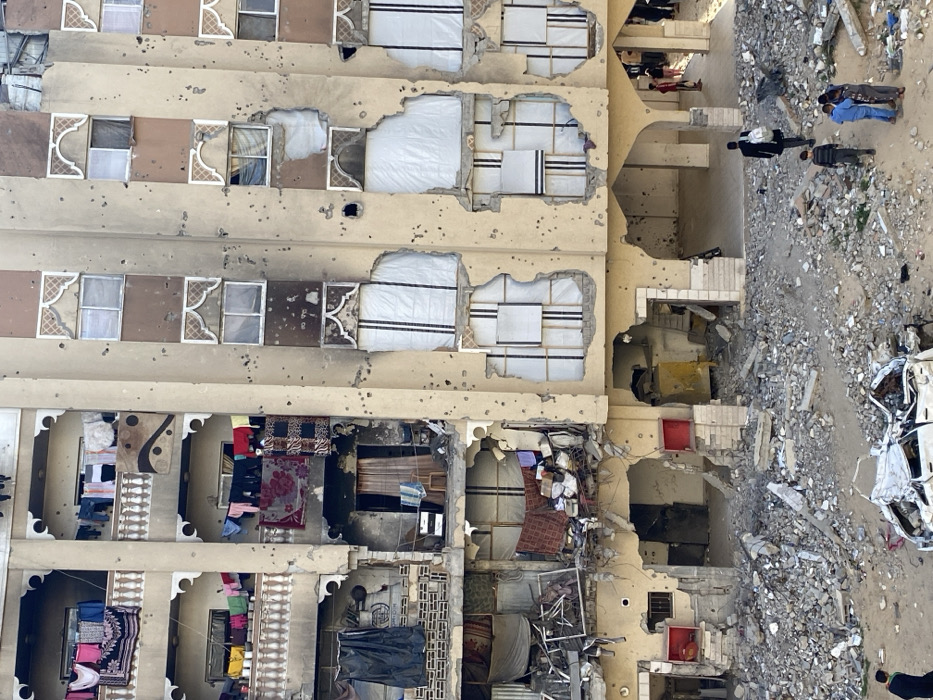This image depicts a multi-story building in Khan Yunis, Palestinian Territory, exhibiting extensive damage. The structure, appearing to be concrete or stucco, features numerous impact holes and bullet marks across its facade.
On the left side, four upper floors show balconies with white balustrades. Laundry, including shirts, blankets, and towels, hangs on clotheslines from these balconies. A large patterned rug is draped over a third-story balcony from the top. Windows on these floors are mostly intact, though some are broken.
The lower three floors on the left side are severely damaged, with large sections of walls and floors collapsed or blown out, exposing wrecked interiors filled with debris and personal items. On the right side, the building facade also shows heavy damage, including broken windows and large impact craters. Several windows on these middle floors are covered with white tarps or sheeting, secured by visible wooden frames, suggesting makeshift repairs. Some window designs feature decorative arched tops.
The ground level is covered in a substantial amount of concrete rubble, rebar, and debris directly in front of the building. A heavily damaged, crumpled white car chassis lies amidst the rubble on the left. Several individuals are present on the ground. A man in a dark suit jacket and pants, carrying a light-colored backpack, walks away from the building towards the right. A group of three children or young individuals stands near him. Additional figures are visible further right, within the arched openings of the ground floor. Another individual in a dark jacket is partially visible on the far left, near the damaged car. The scene is illuminated by daylight.