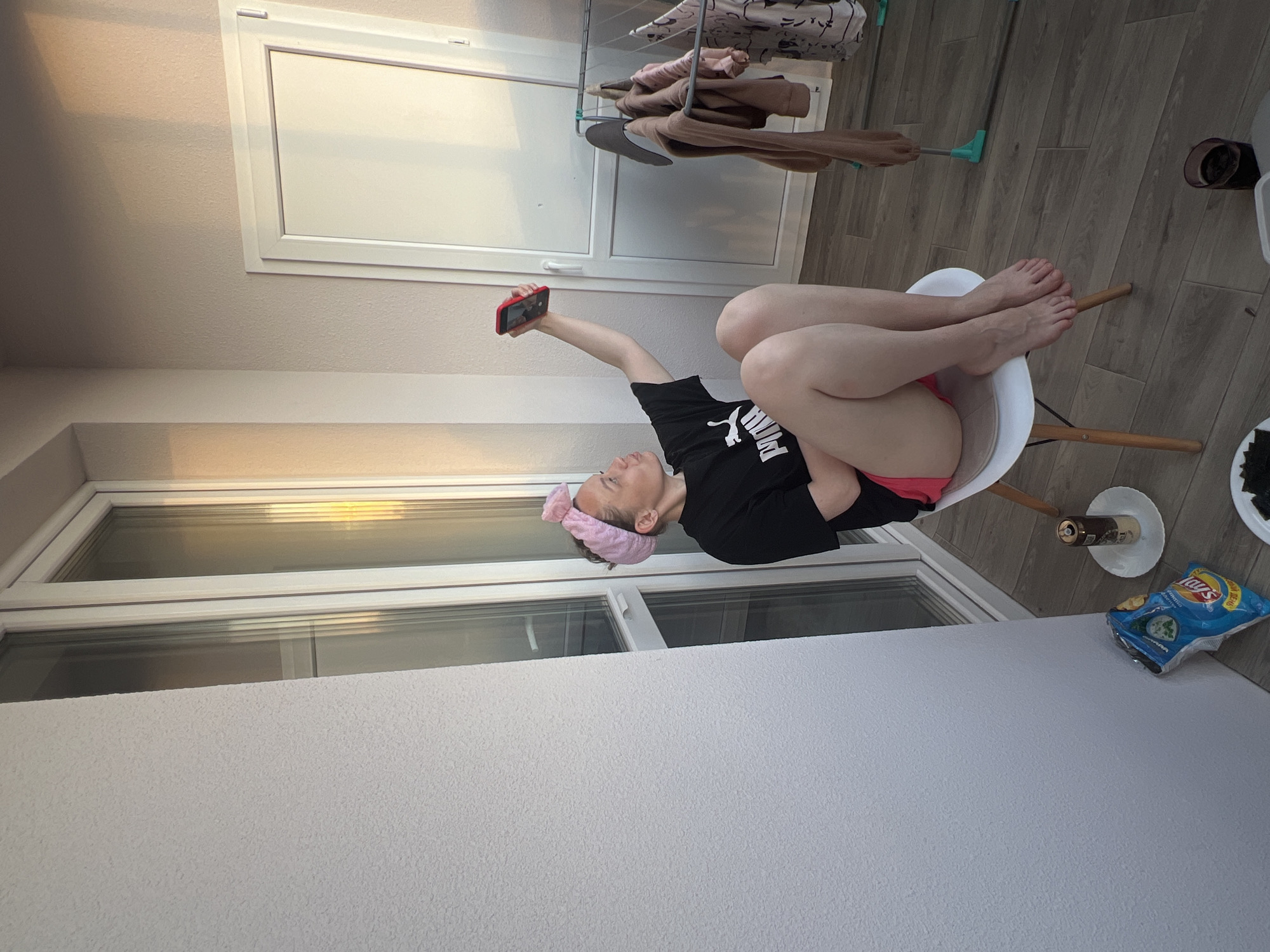 A woman with a pink headband sits on a white chair on a balcony in Izhevsk, Russia, holding a red phone in her outstretched hand, seemingly taking a selfie. She wears a black Puma shirt and what appears to be pink shorts. A bag of Lay's chips sits on the white railing of the balcony. A drink, possibly a beer, rests on a white coaster next to the chips. A drying rack with clothes hangs near a window. The flooring appears to be wood-look tiles.