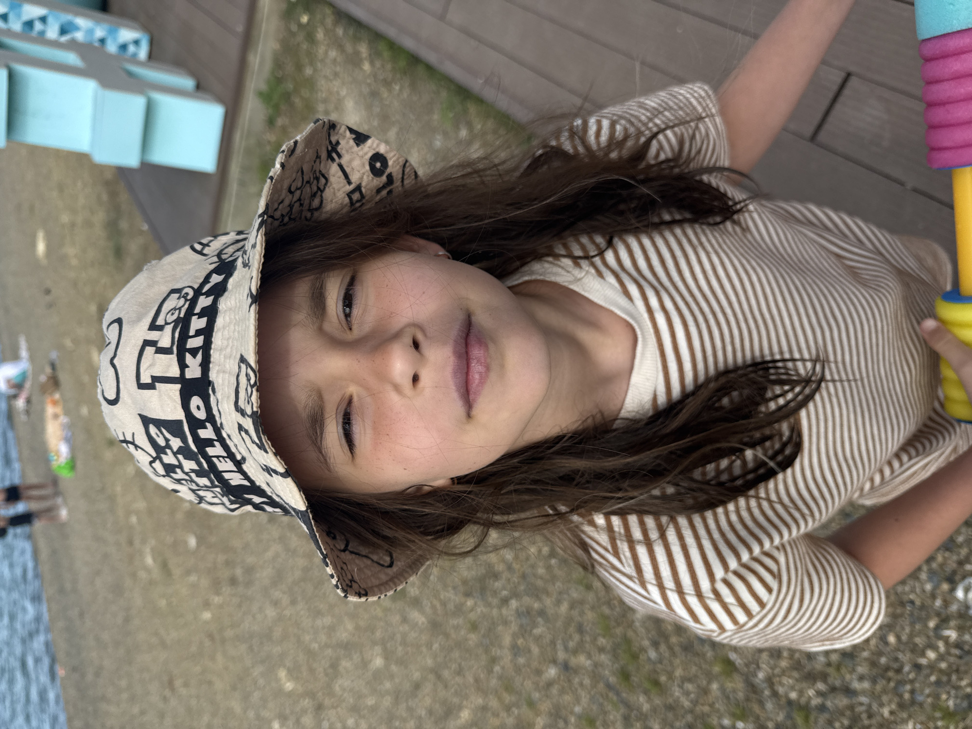 The image features a young girl captured in a portrait-style shot, looking directly at the camera with a gentle, relaxed smile. Her long, dark brown hair cascades around her shoulders. She has a fair complexion with noticeable freckles across her nose and cheeks.
The girl is wearing a light-colored bucket hat adorned with black patterns. Distinct text visible on the hat includes the English phrase "MELLO K" and what appear to be Japanese characters, such as "ーロ". She is dressed in a light-colored, possibly off-white, long-sleeved shirt with thin, vertical brown stripes. In her right hand, visible on the right side of the frame, she holds a colorful water toy composed of stacked rings in pink, light blue, and yellow, attached to a yellow handle.
The setting appears to be an outdoor, possibly recreational area during daylight hours. To the far left, a body of water is visible, suggesting a lakeside or seaside location, with blurry figures standing nearby. The ground beneath her is a mix of light-colored pebbles or sand. In the background, there are light blue, block-like structures, possibly seating or play equipment, and a wooden deck or walkway. The overall atmosphere is bright and casual. The image is rotated, presenting the scene from a slightly tilted perspective.
