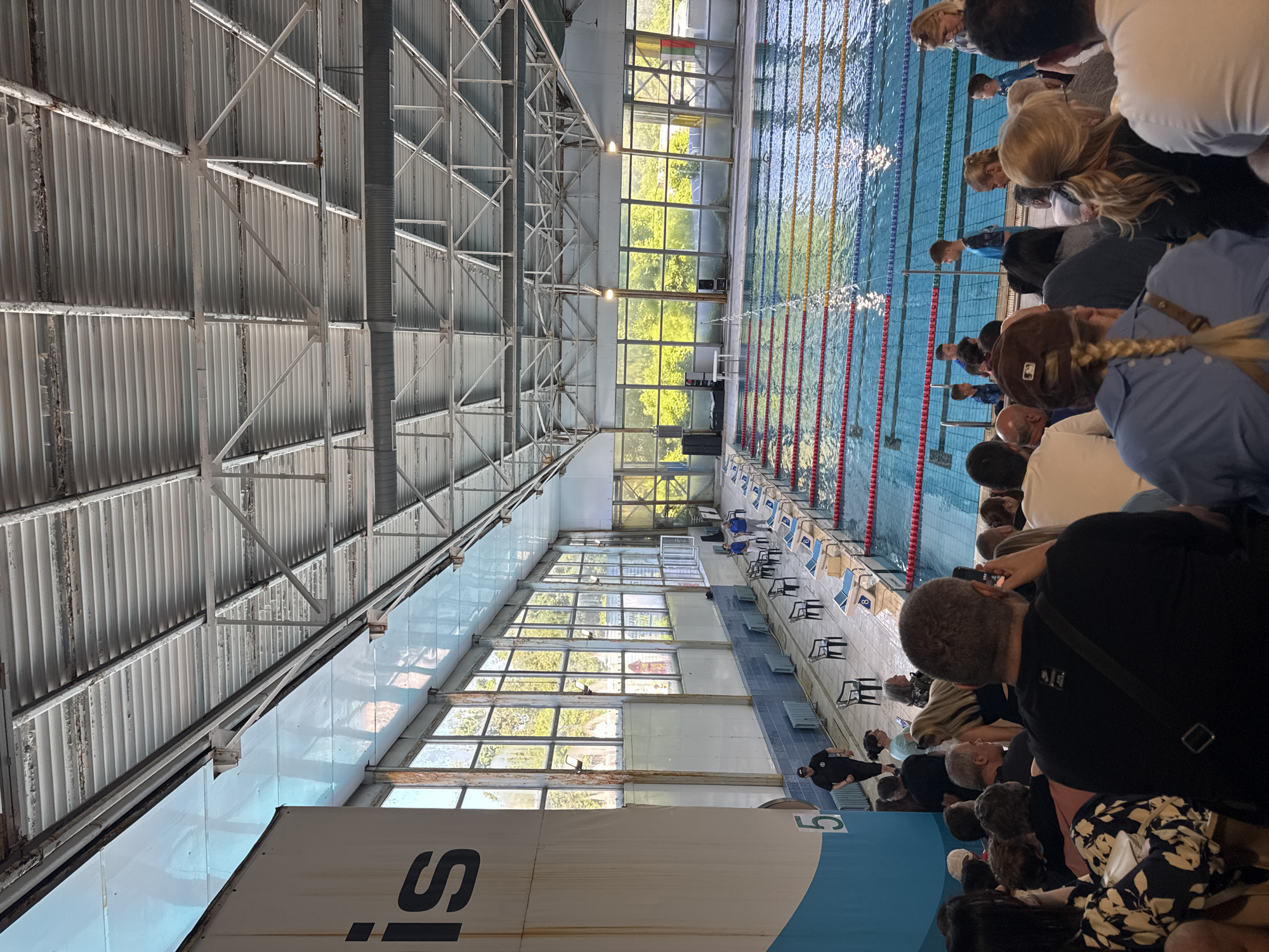 The image shows an indoor swimming pool in Sofia, Bulgaria, viewed from an elevated perspective. The pool has multiple lanes marked with floating dividers. At the far end, starting blocks are positioned. Spectators are seated along the side of the pool, some of whom are watching the activity in the pool. The pool building has a high ceiling with exposed metal structural supports. Large windows provide natural light, and trees can be seen outside. A large white banner is visible near the camera, with the letters "is" partially visible. People are wearing casual clothing.