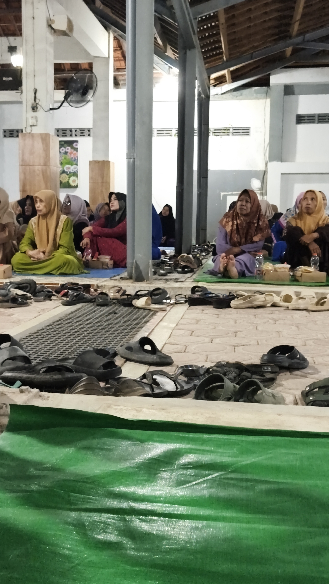 The image depicts an indoor gathering of Muslim women in Dalegan, Indonesia. The women, wearing hijabs and modest attire, are seated on the floor, some on blue and green mats. They appear to be listening or observing something, with varying expressions of engagement. Scattered around them are numerous pairs of sandals and flip-flops, suggesting a place of worship or a communal gathering where shoes are removed. The environment appears to be a simple building with exposed ceiling beams and industrial-style pillars. A green tarp covers the foreground, obscuring the lower part of the scene. In the background, a fan is mounted on the wall, and decorative artwork is visible. The lighting is somewhat dim, creating a subdued atmosphere.