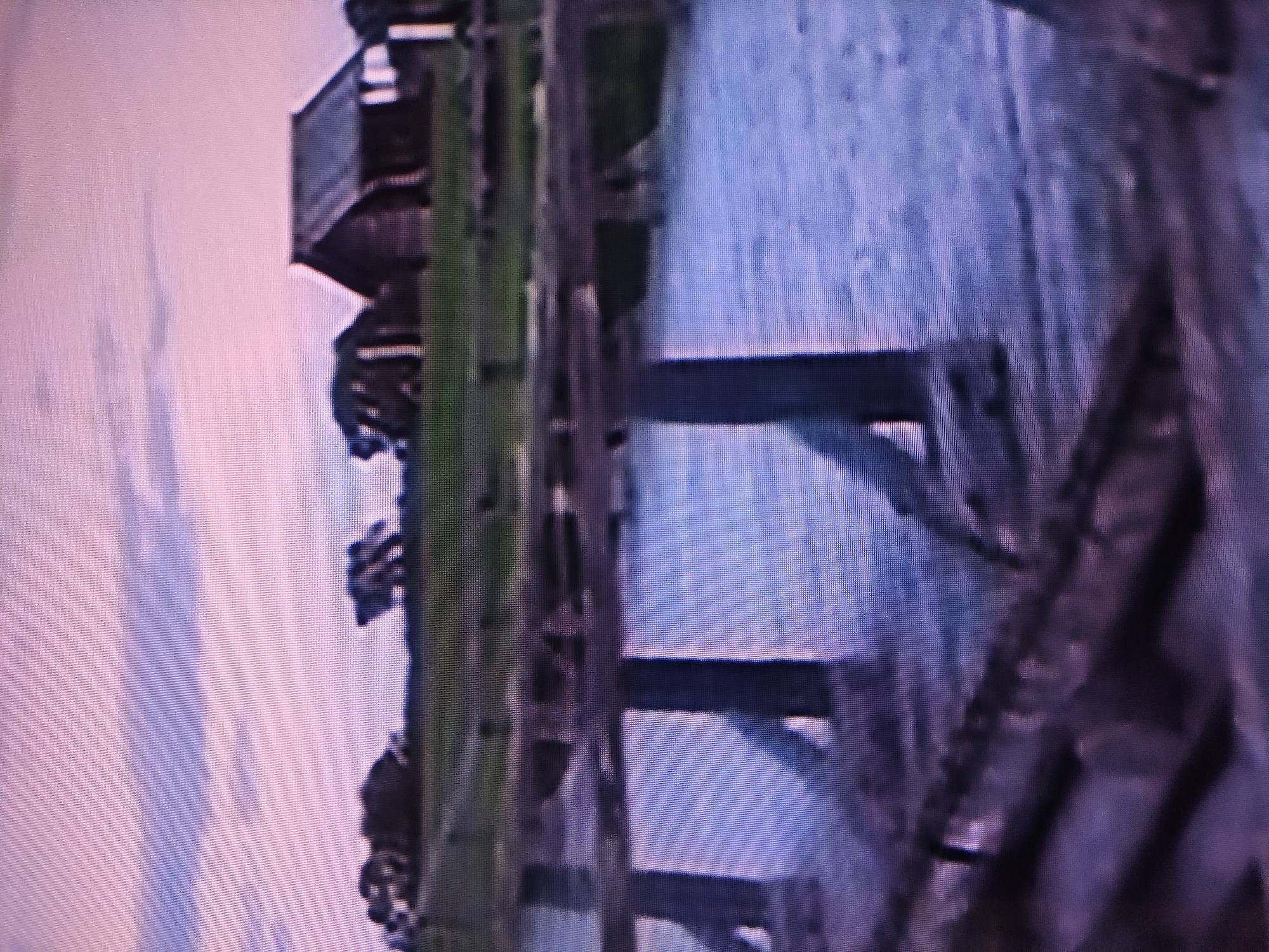 The image shows a waterfront area in Pavlodar, Kazakhstan. A structure, possibly a viewing platform or small building, is visible along the waterfront. Wooden supports or beams extend over the water. The sky is cloudy or overcast.