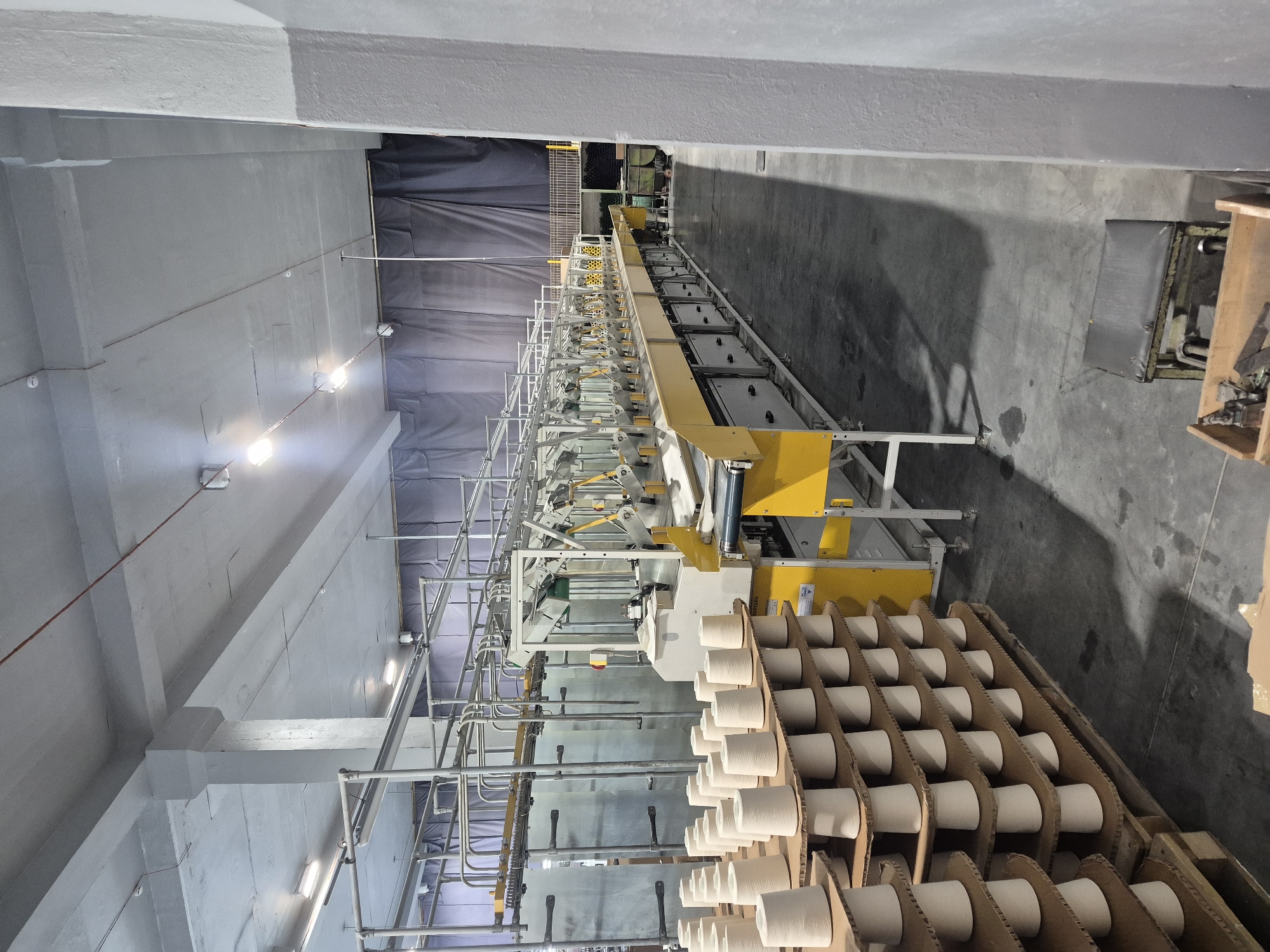 The image depicts a textile manufacturing facility, likely in Chaikovsky, Russia, based on context. A long, yellow and white automated spinning machine dominates the scene, stretching into the background, with a large curtain partially obscuring the far end of the room. In the foreground, a pallet holds cardboard trays filled with numerous spools of white yarn. The room is industrial, with a gray concrete floor and ceiling, illuminated by evenly spaced overhead lights. No people are visible in the image.