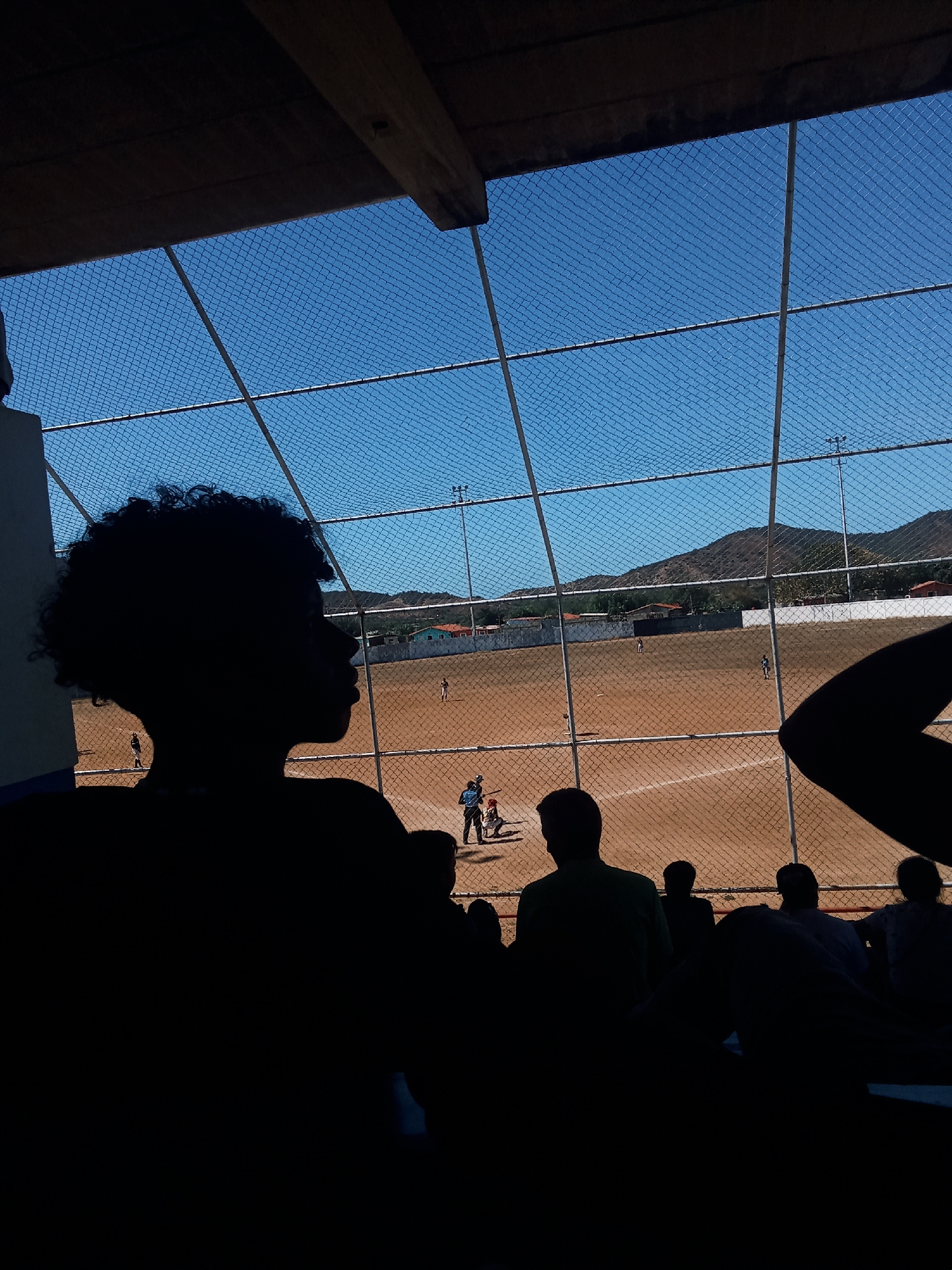 The image depicts a baseball game in Juan Griego, Venezuela, viewed through a chain-link fence. Silhouetted figures of spectators, including a person with curly hair in profile, are in the foreground, sitting in stadium seating under a covered structure. Beyond the fence is a dirt baseball field with players in motion. In the background, there are buildings and low mountains under a clear blue sky. Light poles are visible around the perimeter of the field.