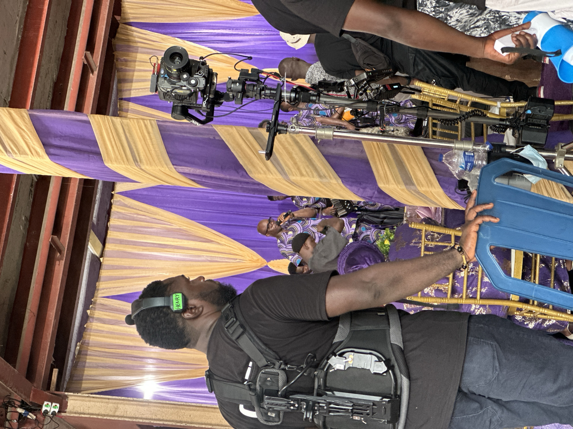 A filming scene is set under a tent with alternating purple and gold fabric stripes. A man wearing a black shirt and jeans is facing away from the camera with a stabilizing rig strapped to his body and headphones on his head. A black movie camera mounted on a tripod is visible overhead, pointed toward the scene. Other individuals in patterned clothing are positioned in the background, some appearing to be musicians. Another person in black stands near the camera equipment. A blue plastic item is held in the lower right corner. The presence of electrical outlets and exposed beams suggest the tent may be an improvised setup. The location is Lagos, Nigeria.