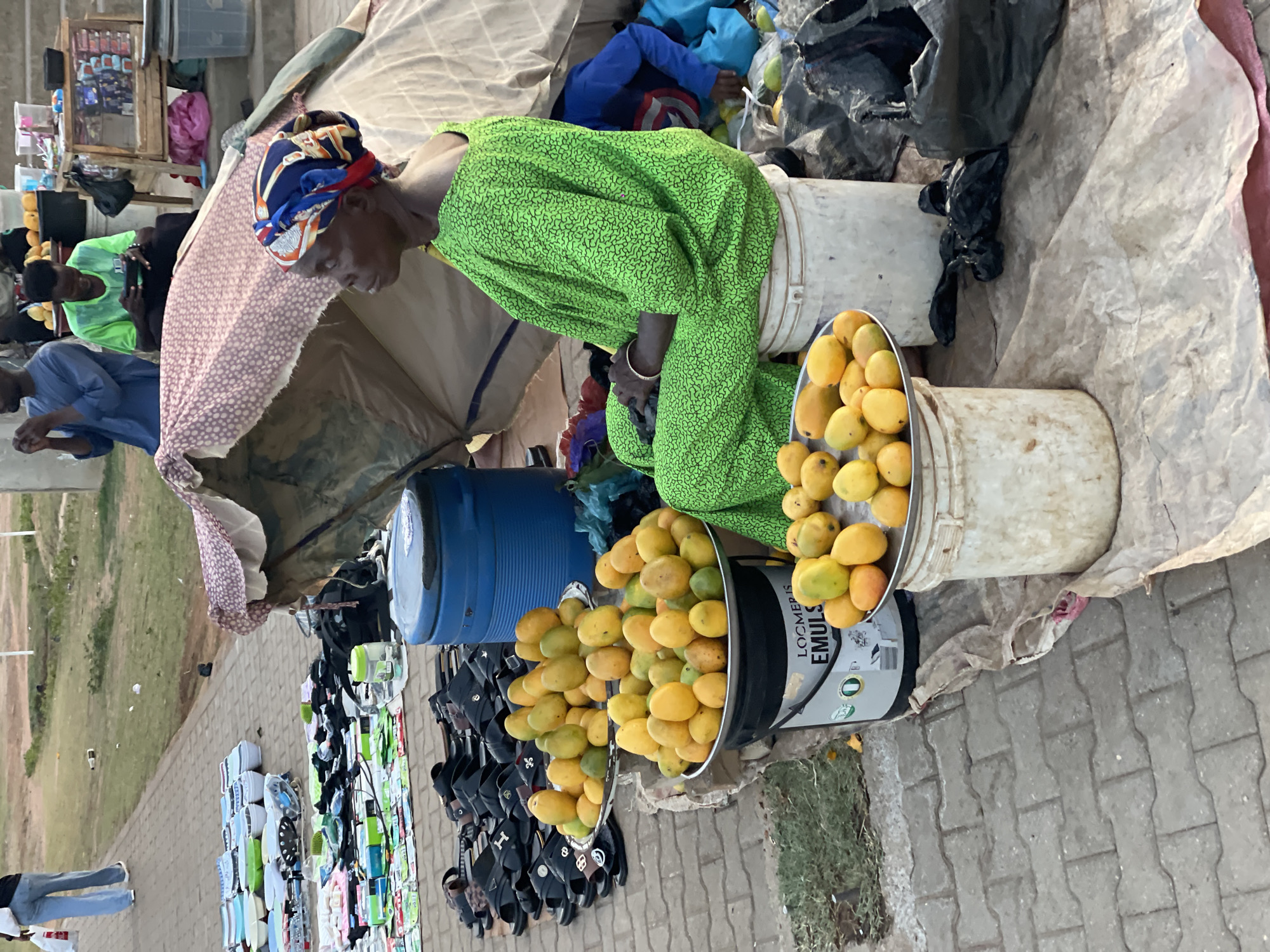 A woman in a green patterned dress and colorful head wrap sits next to two buckets overflowing with yellow mangoes at an outdoor market stall. The stall is set up under a makeshift shelter made of fabric draped over a frame, with a blue barrel visible nearby. Various items, including footwear, are displayed on the ground. The setting appears to be Galadimawa Village, Nigeria, indicated by the style of clothing and goods.
